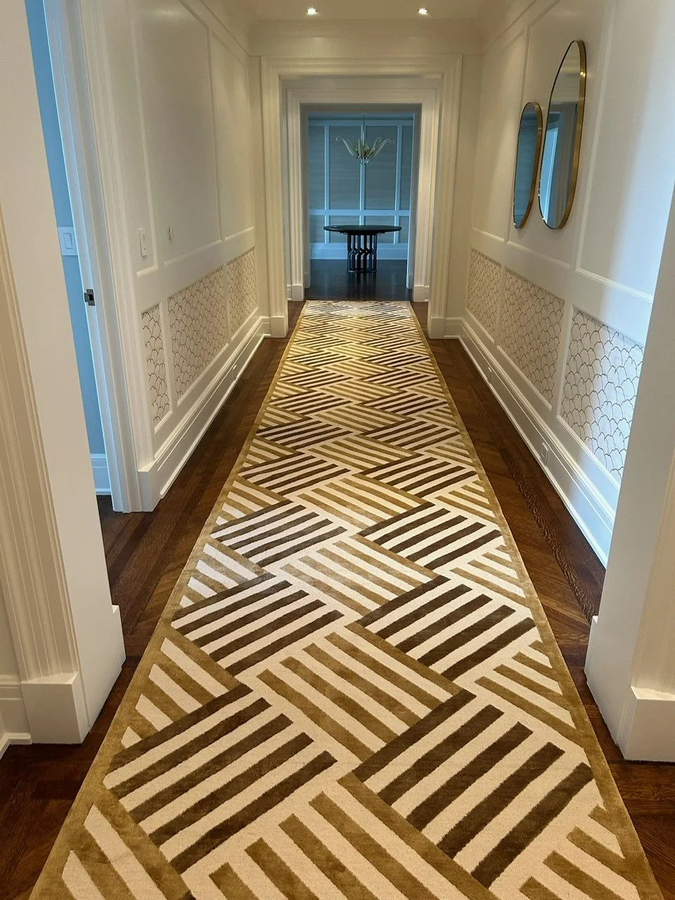 A hallway with a patterned runner rug, white walls with decorative wall panels, and two round mirrors on the right wall. The hallway leads to a room with a dark table and a chandelier.