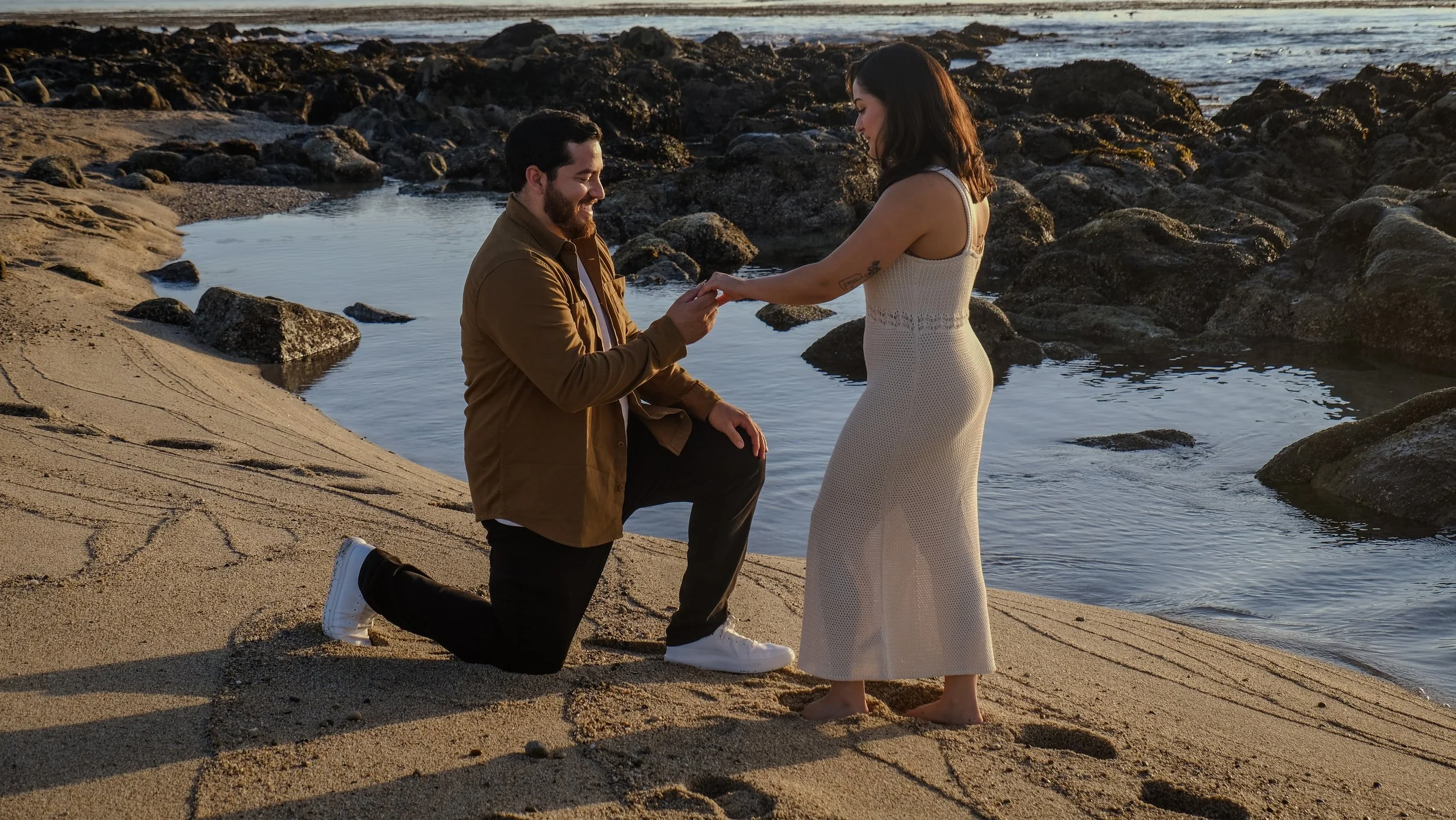 A man proposing to a woman on a sandy beach with rocks and water in the background at sunset.