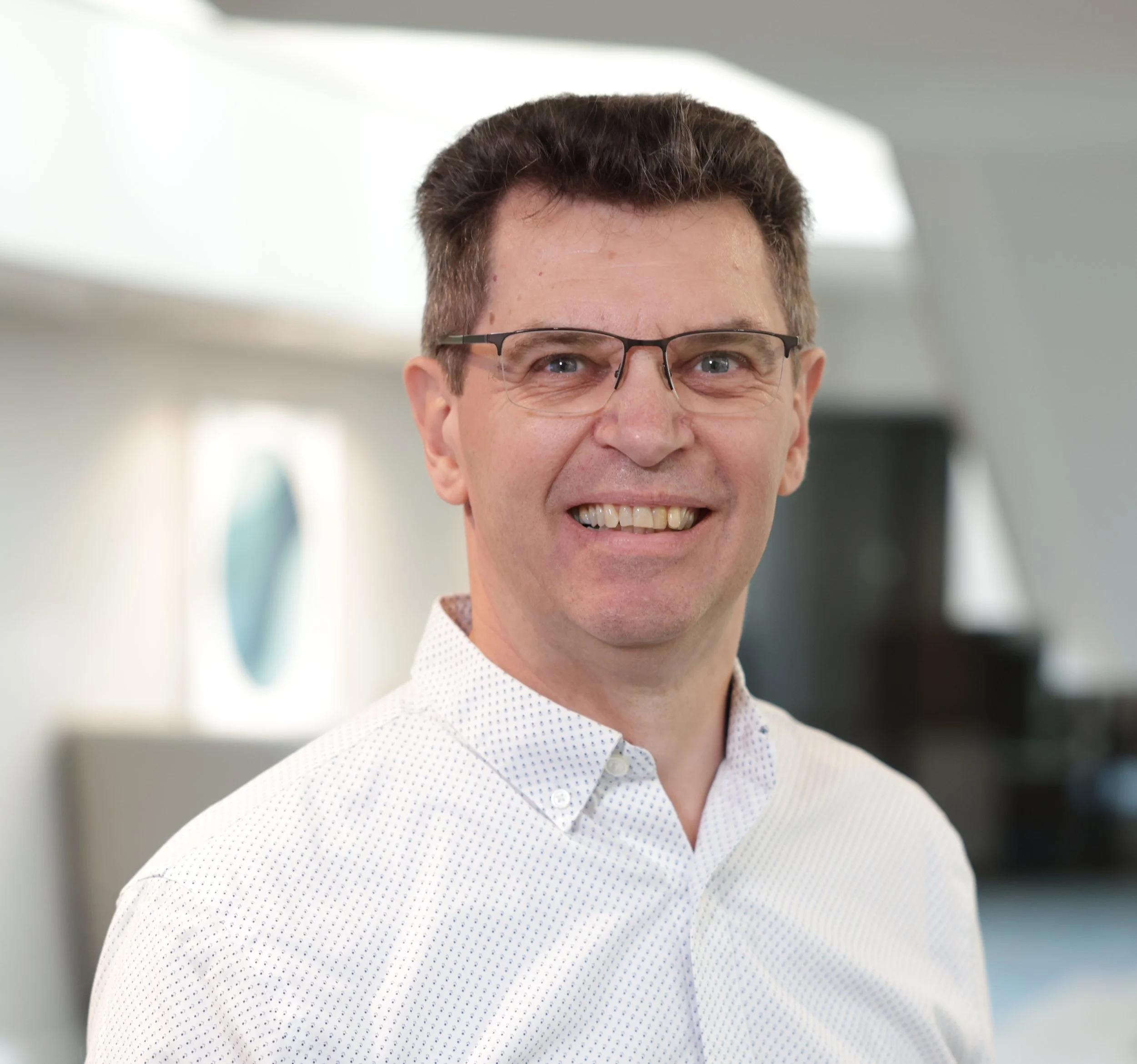 A man with glasses speaking into a microphone, wearing a light-colored blazer and patterned shirt.