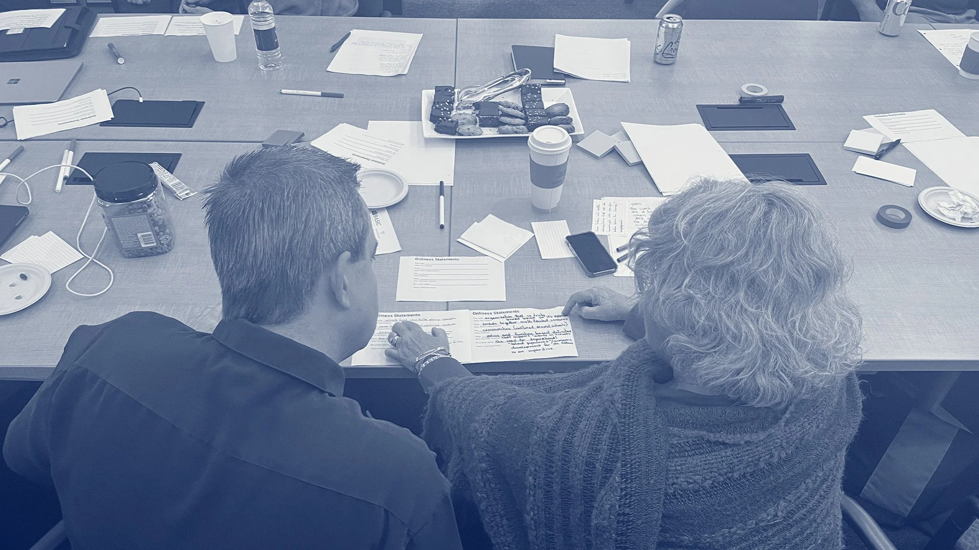 A man and a woman sit beside each other at a table reviewing a handwritten note. Around them, the table is scattered with pens, notebooks and beverages—evidence of a large group collaboration