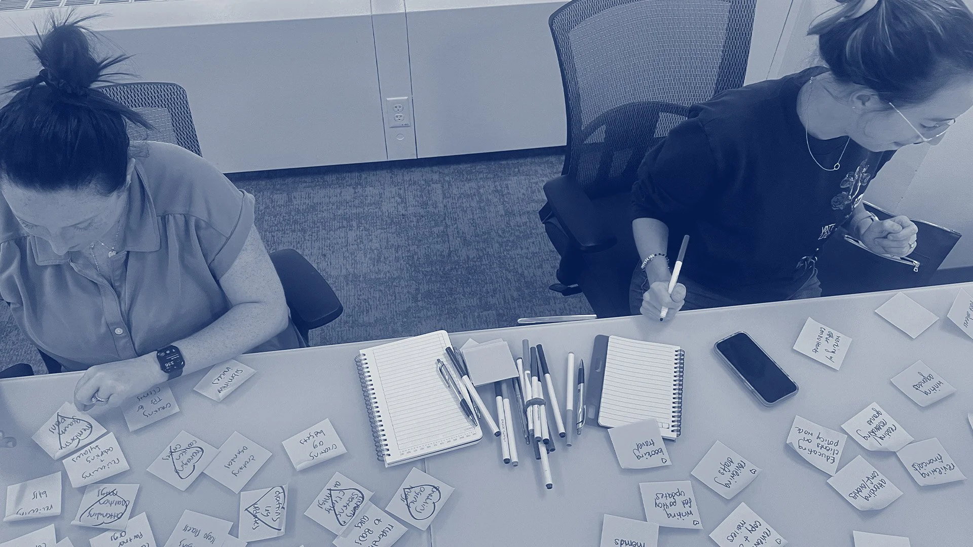 Two women sitting at a conference table scattered with notebooks, sticky notes and pens