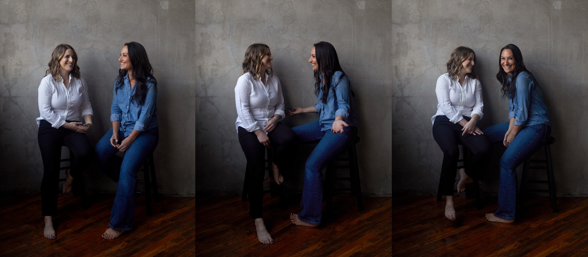 Two women sitting on chairs and interacting, one wearing a white shirt and black pants, the other in a blue denim shirt and jeans, smiling and engaged in conversation, against a plain gray wall and wooden floor.