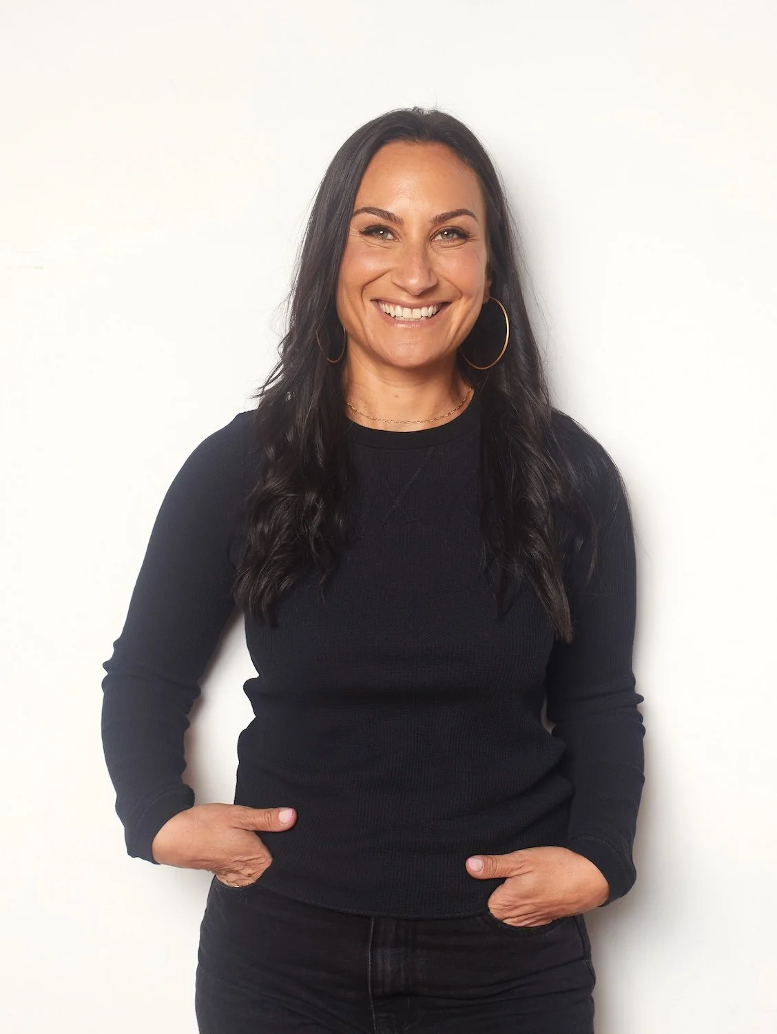 A portrait Jenna Shapiro, wearing black jeans and a black shirt, standing against a white wall