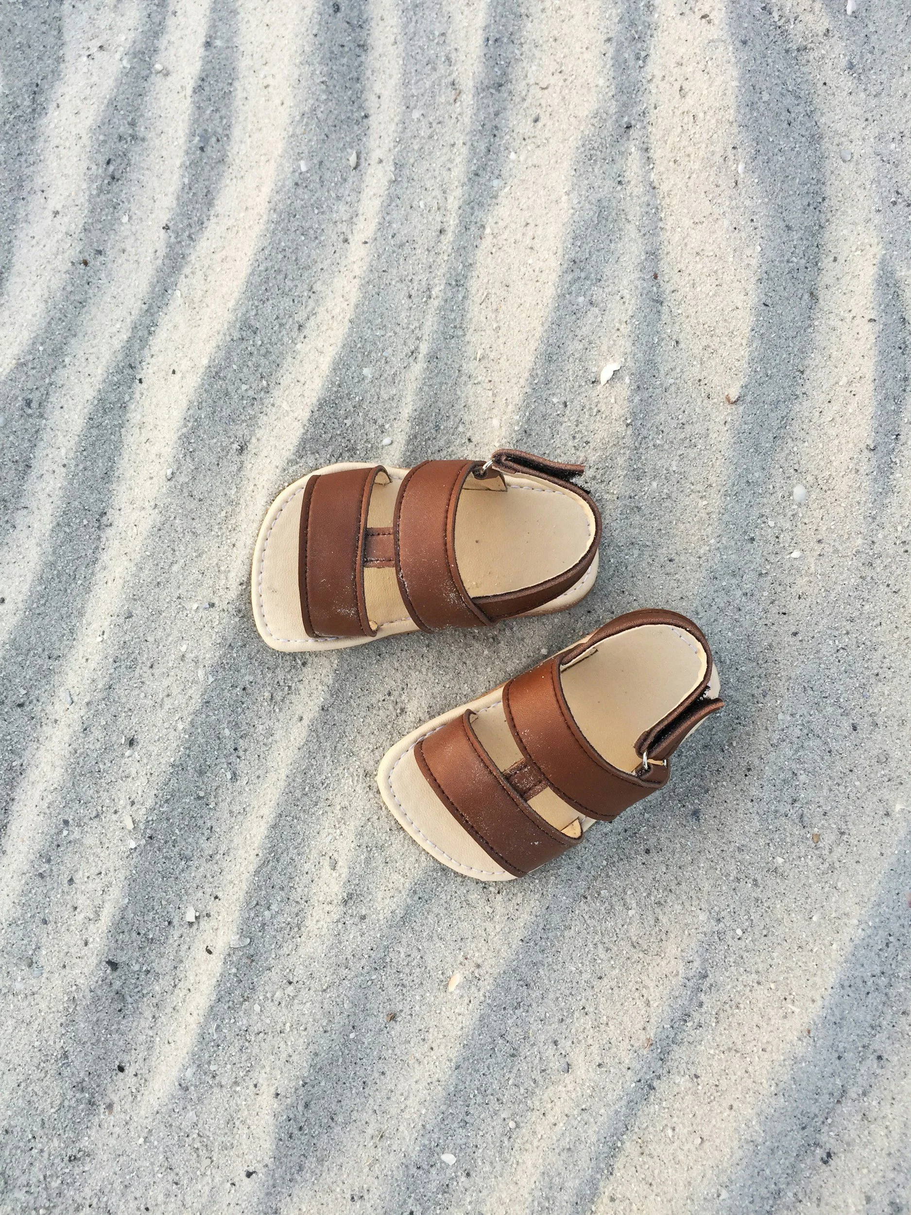 A pair of brown children's sandals on sandy ground with rippled texture.