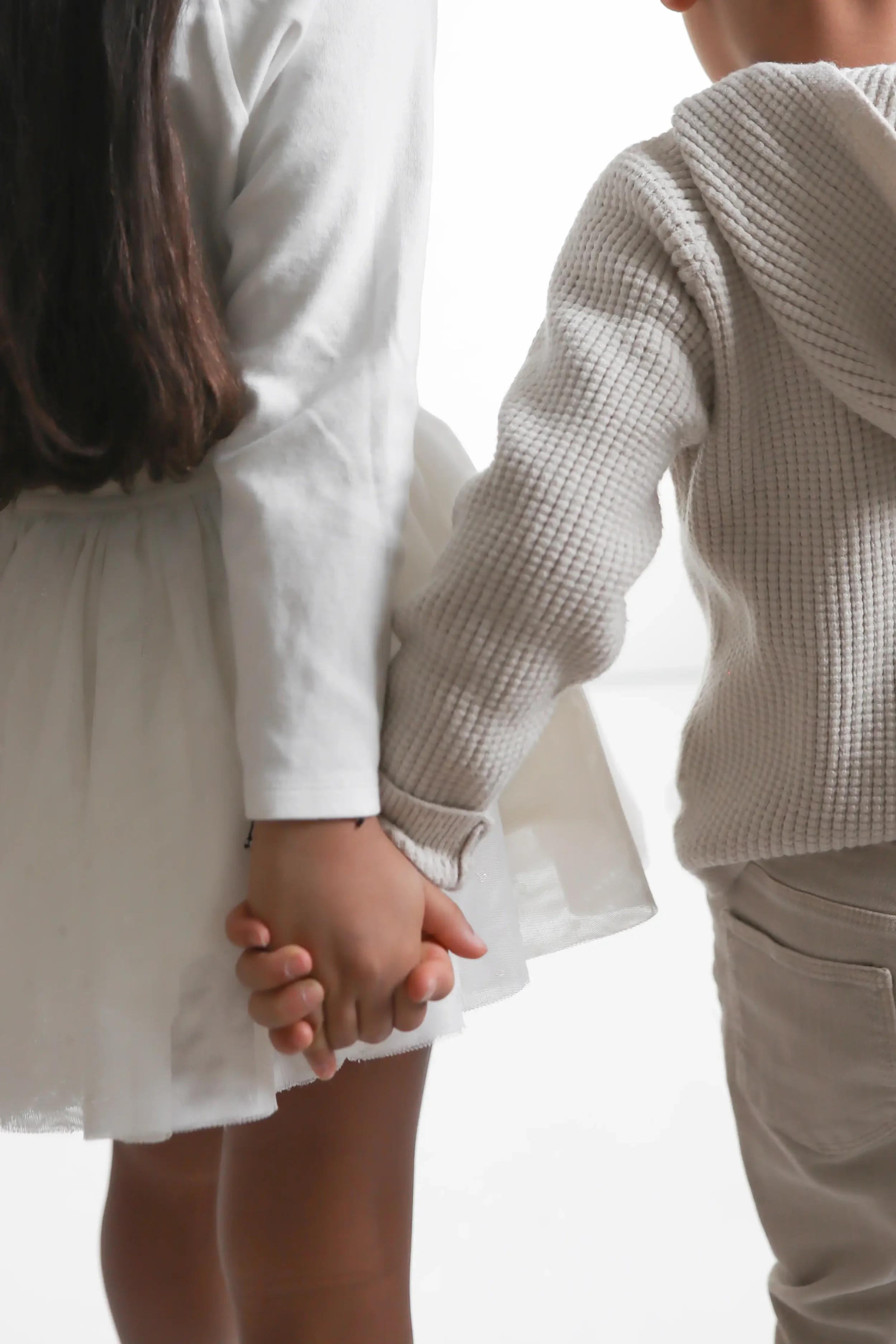 A close-up of a girl and boy holding hands, with the girl wearing a white dress and the boy wearing a beige textured sweater.