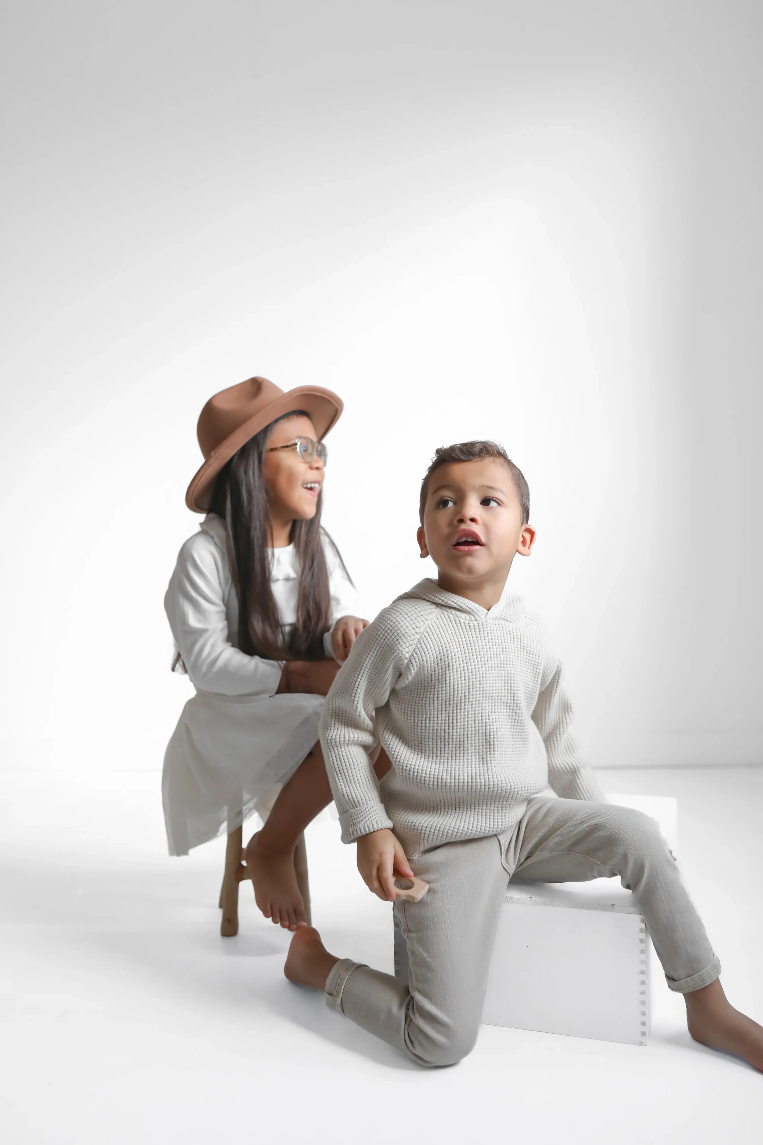 A girl with glasses and a brown hat sitting on a stool, smiling and holding the shoulders of a young boy who is kneeling on the floor with a neutral expression, both wearing light-colored clothing against a blank white background.