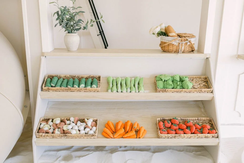 Miniature vegetable and fruit models on wicker trays, including green peas, celery, green peppers, white beans, carrots, and strawberries, arranged on a white wooden shelf with a small plant and a basket of bread in the background.