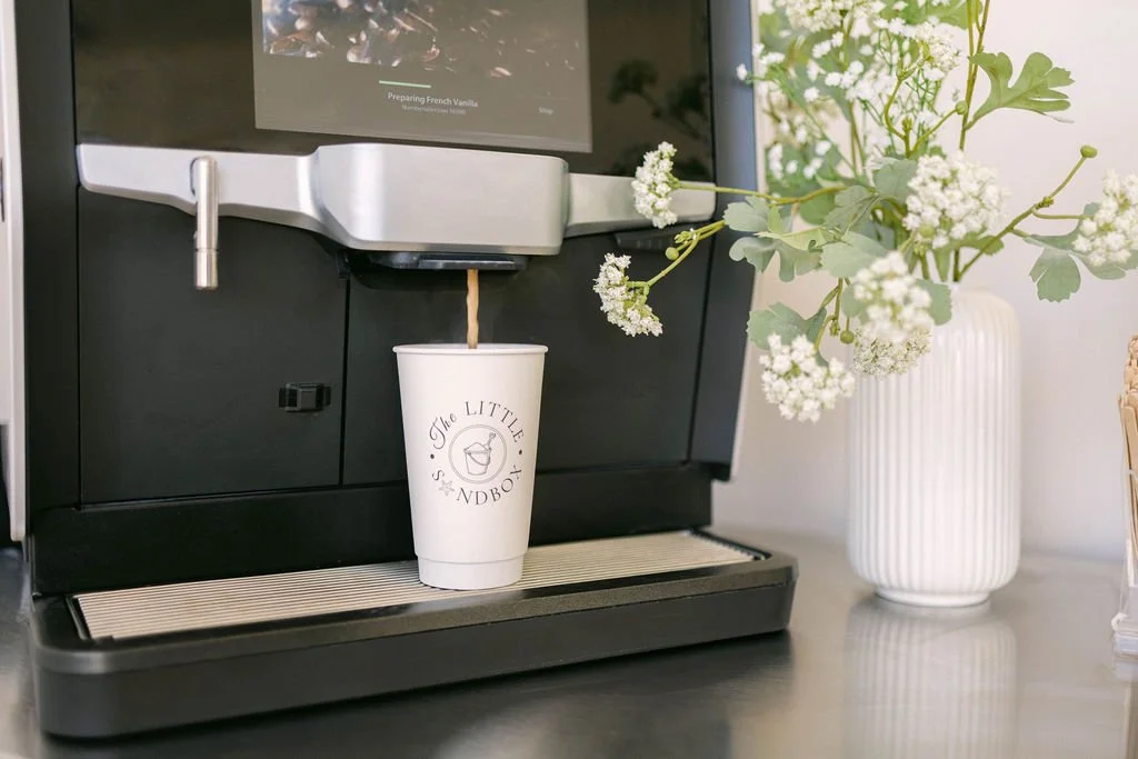 A coffee machine dispensing coffee into a white paper cup labeled 'The Little Sandbox,' with a vase of white flowers nearby.