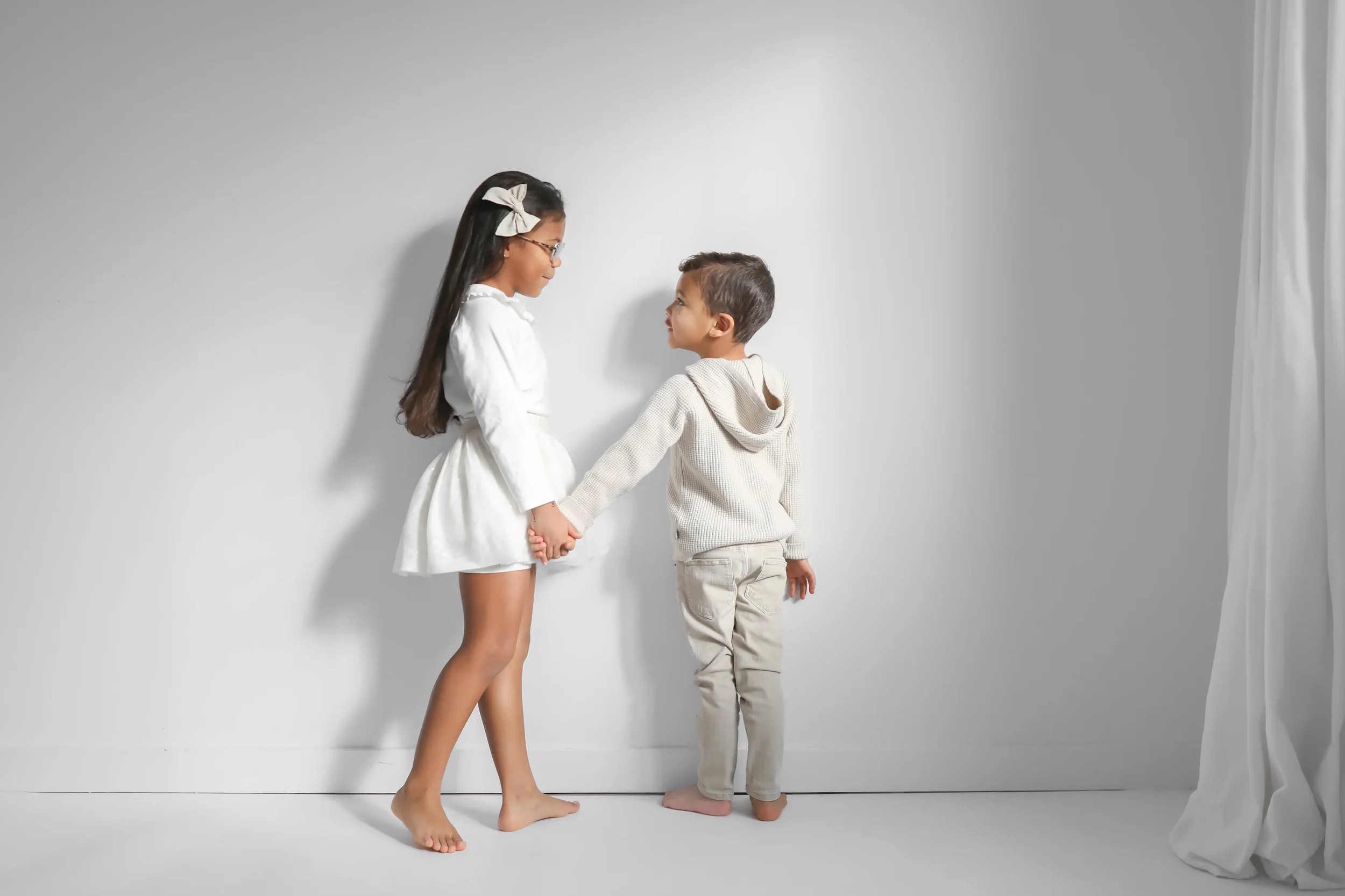 A young girl and boy holding hands and looking at each other in a white room with a white curtain on the side.