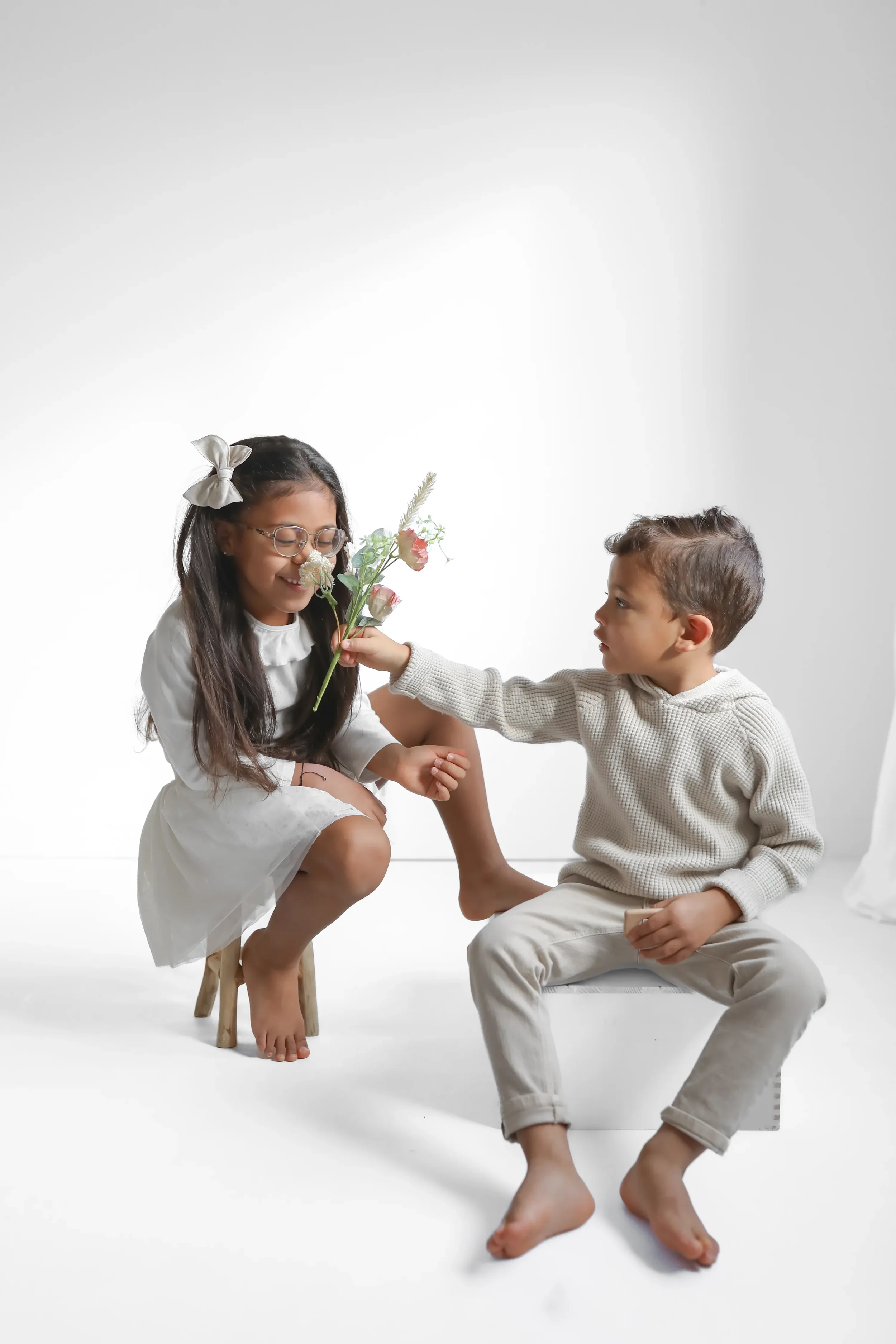 A girl in a white dress with glasses sitting on a small stool, smiling as a boy in a beige sweater and pants hands her a bouquet of flowers while sitting on a white box. The background is white.