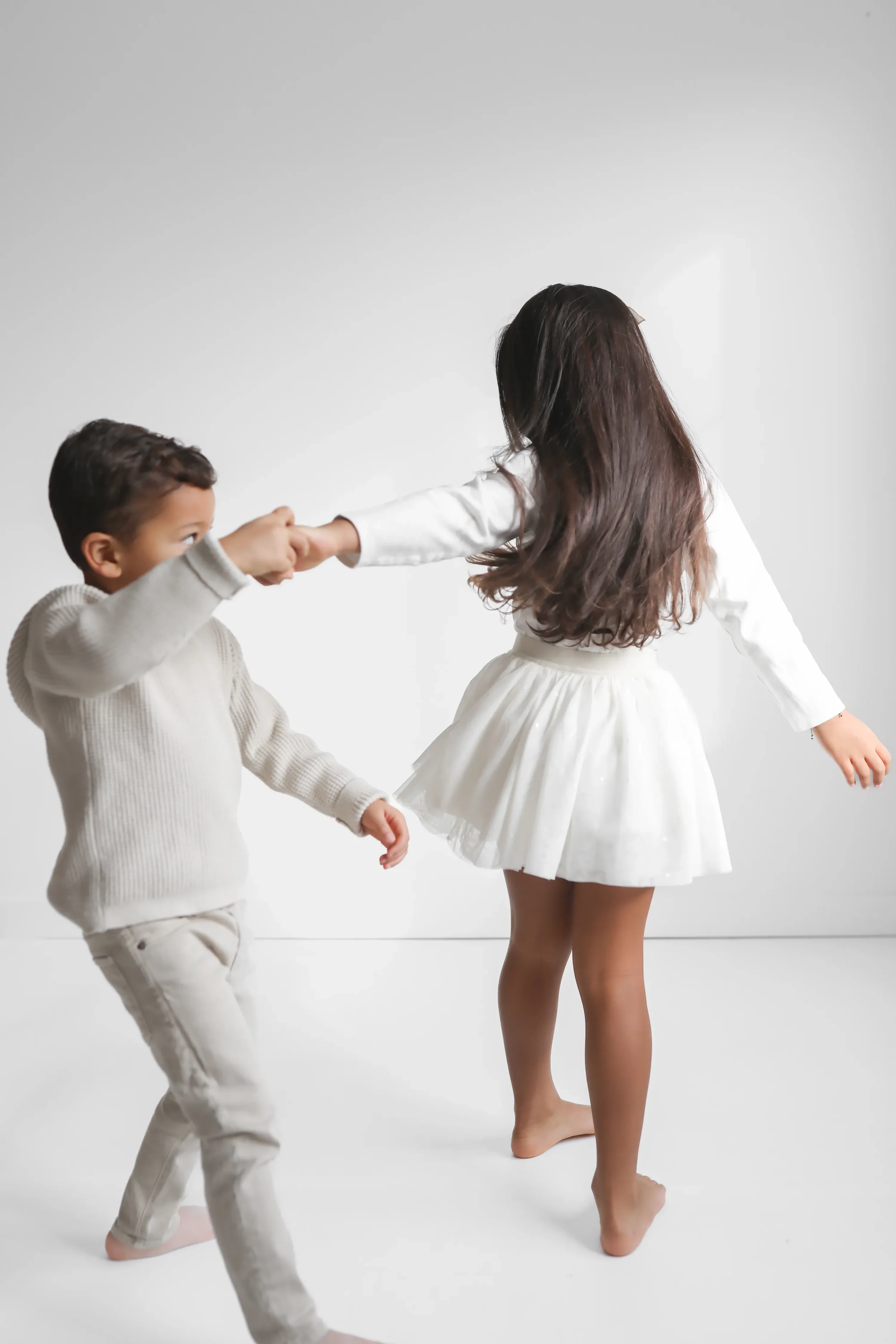 Two children, a boy and a girl, are playing together on a plain white background. The girl is dressed in a white dress and is holding the boy's hand, who is wearing a cream sweater and cream pants. They appear to be spinning or dancing.