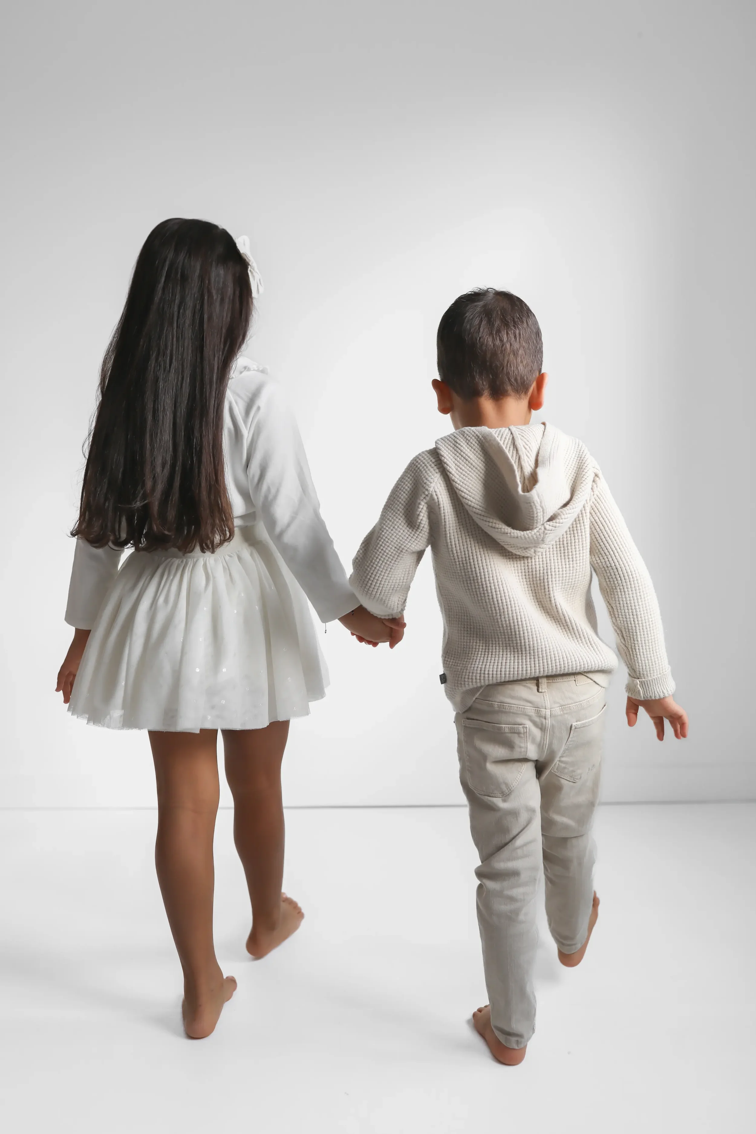 Two children, a girl in a white dress and a boy in a beige hoodie and pants, holding hands as they walk barefoot on a white background.