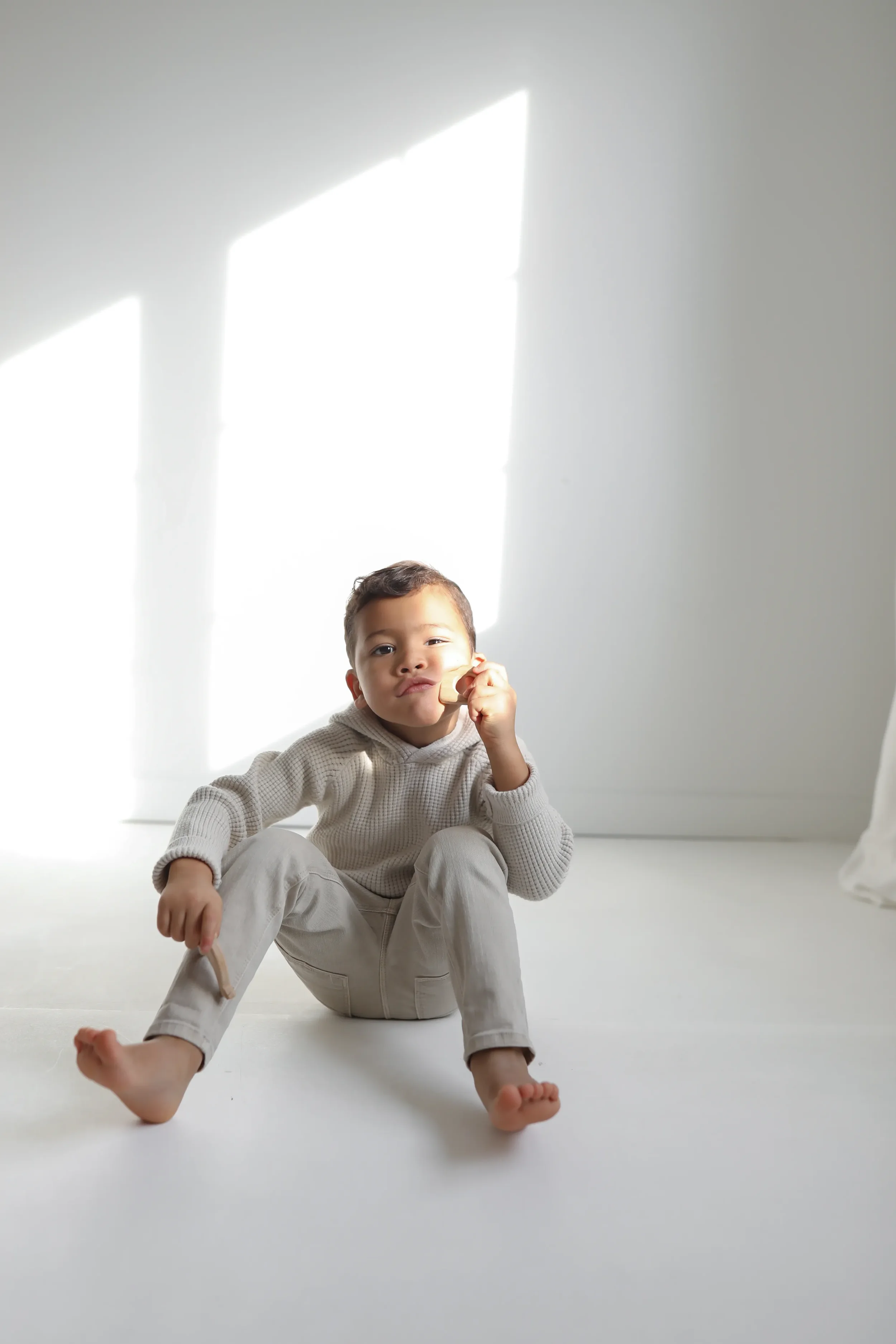Child sitting on a white floor in a bright room with sunlight streaming through a window, wearing a cream-colored hoodie and pants.