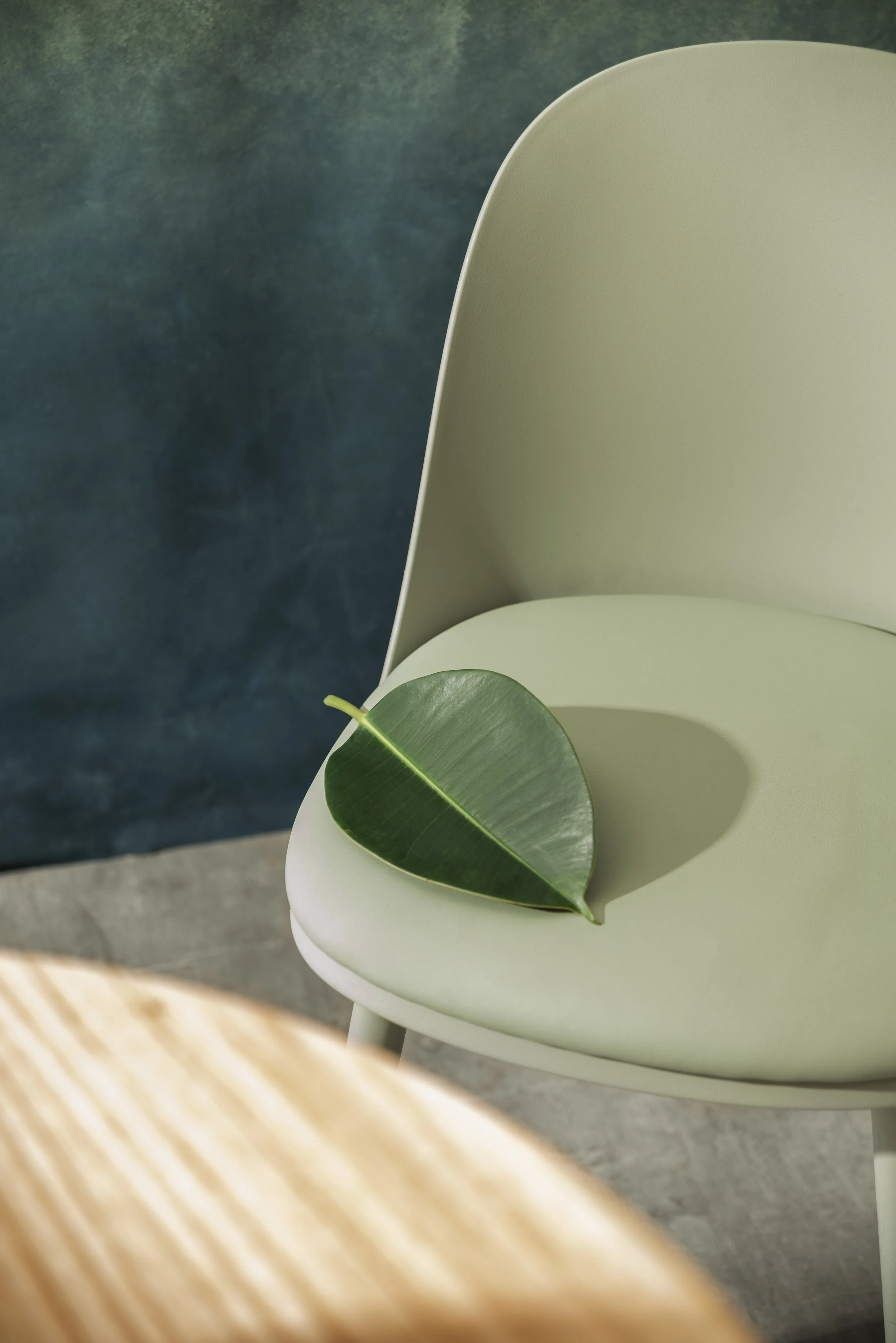 A light green chair with a single green leaf resting on its seat, with a dark blue wall and a wooden table in the foreground.