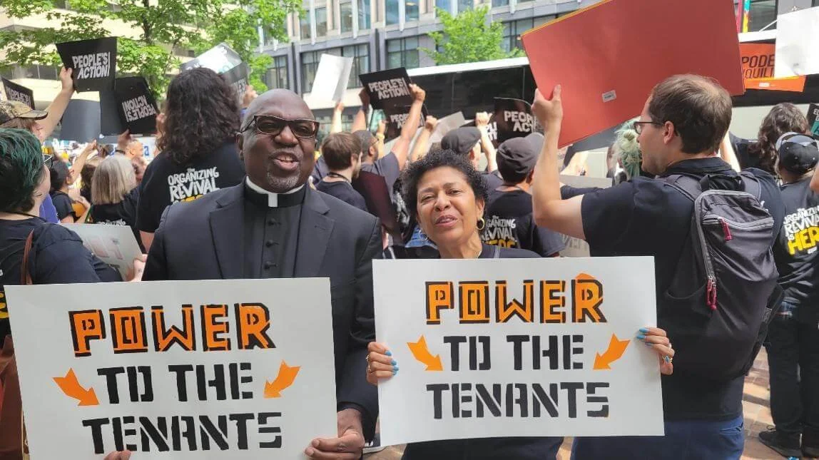 People participating in a protest or rally, holding signs that say 'Power to the Tenant' and 'Organizing Revival'. In the foreground, a man in clerical attire and sunglasses, and a woman, are smiling at the camera.