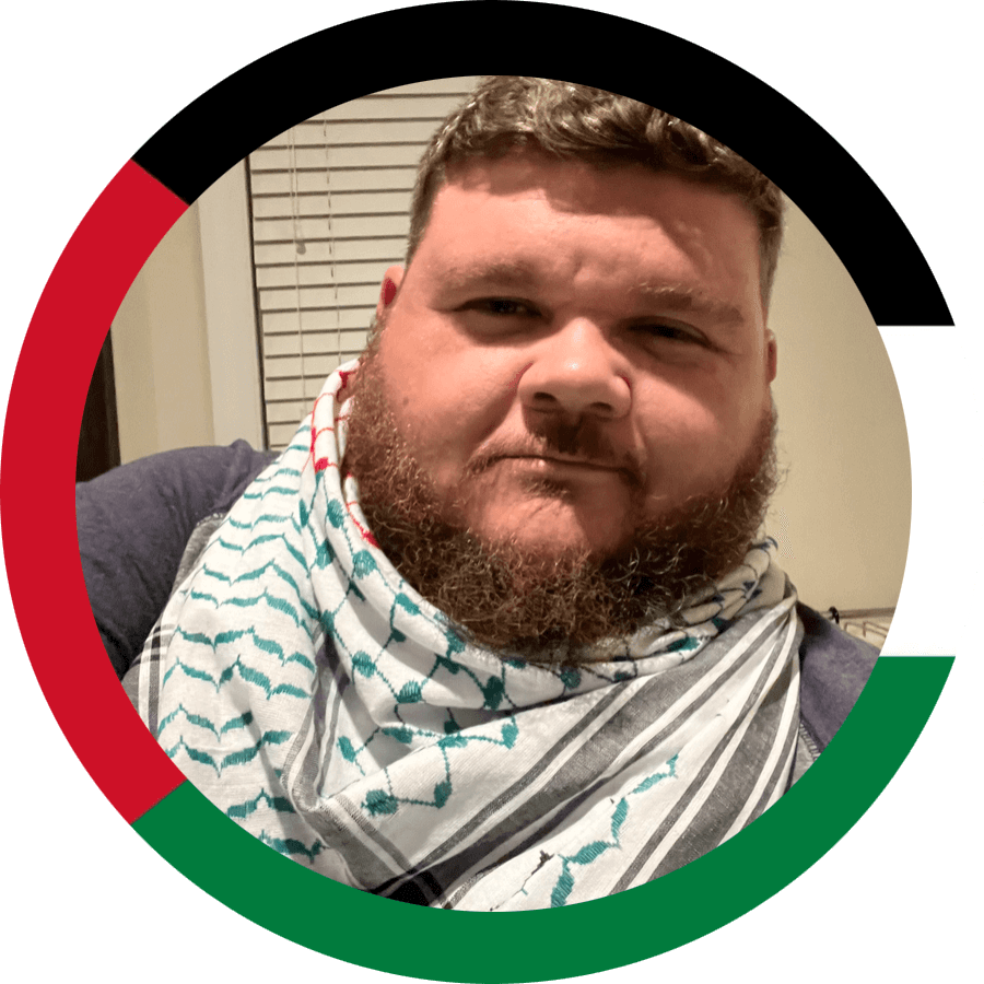 A man with a beard and short hair taking a selfie indoors, wearing a patterned bandana around his neck, with a window with blinds in the background.