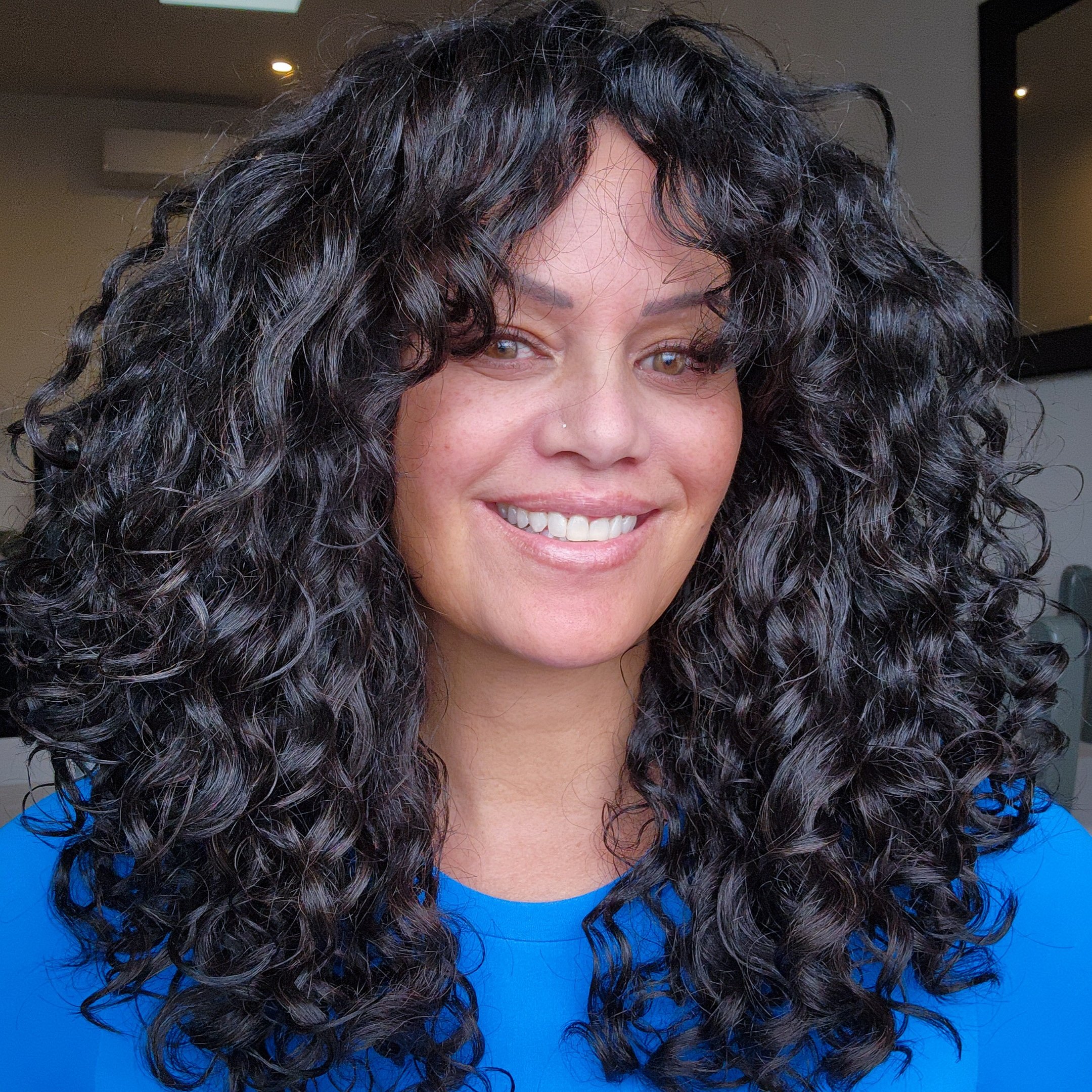 A woman with long, dark, curly hair smiling in an indoor setting.