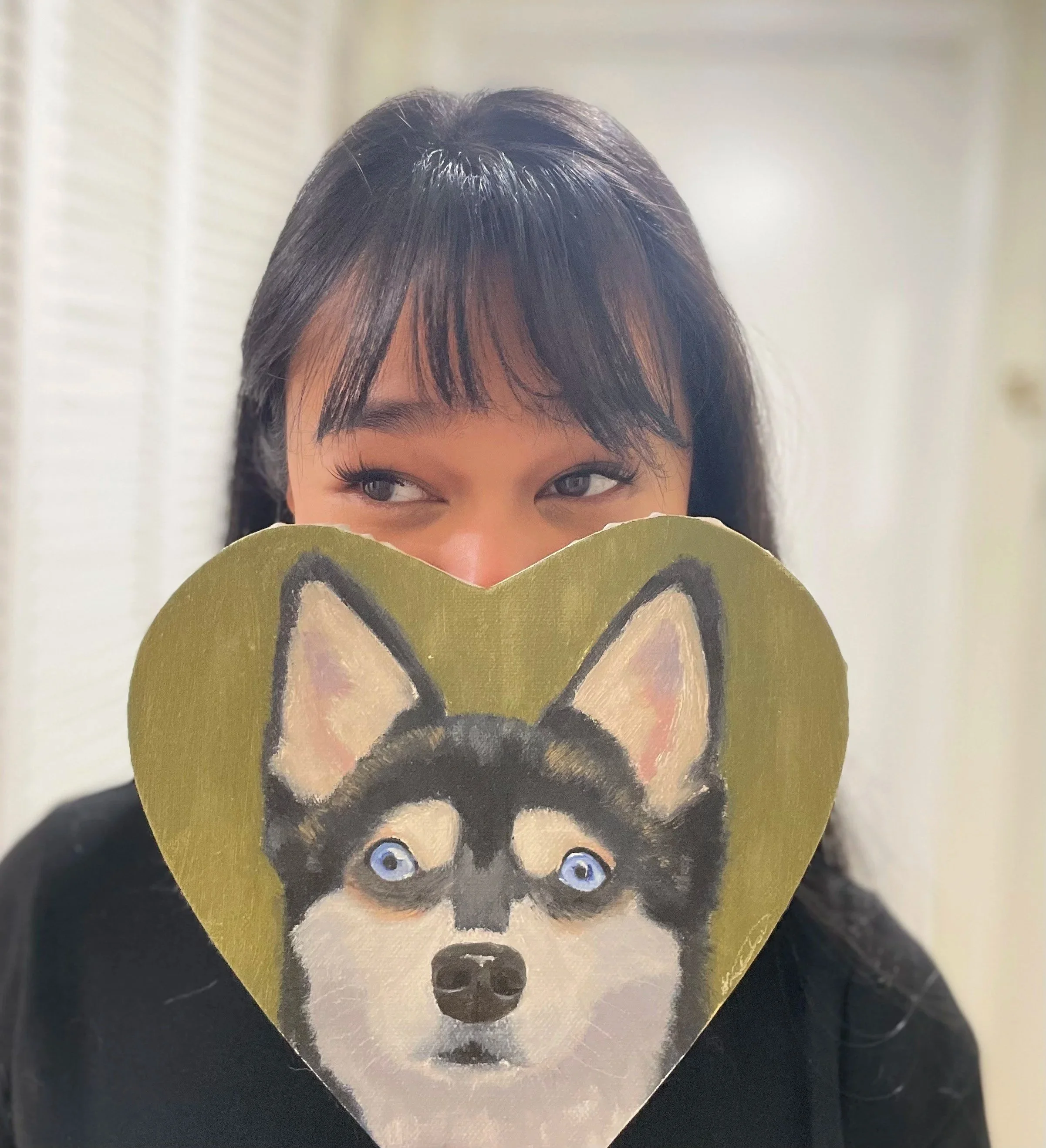 Woman holding a heart-shaped board with a painted Husky dog's face covering her mouth, showing only her eyes and forehead.