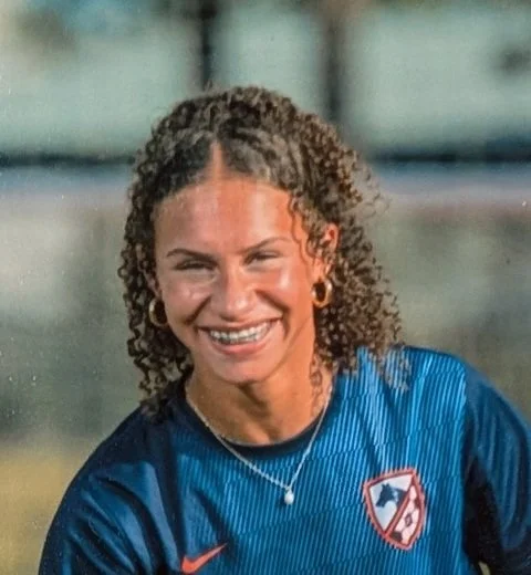 Close-up of a smiling woman with curly hair wearing a blue sports jersey with a logo.