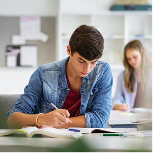 A young man sitting at a desk, studying with open books and writing instruments, with a young woman in the background also studying.