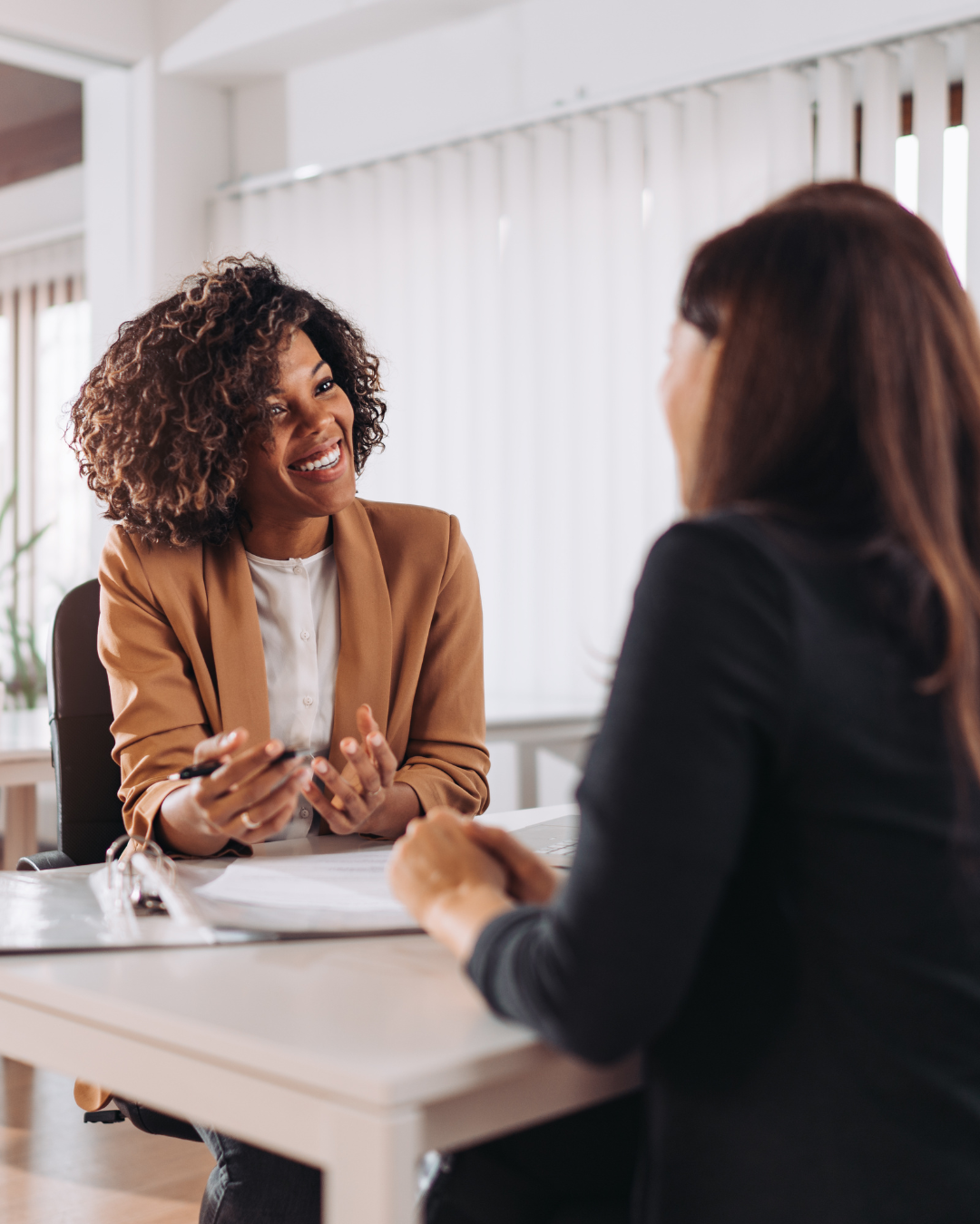 Two women having a conversation in an office, one is smiling and holding a pen, facing the other woman with brown hair.