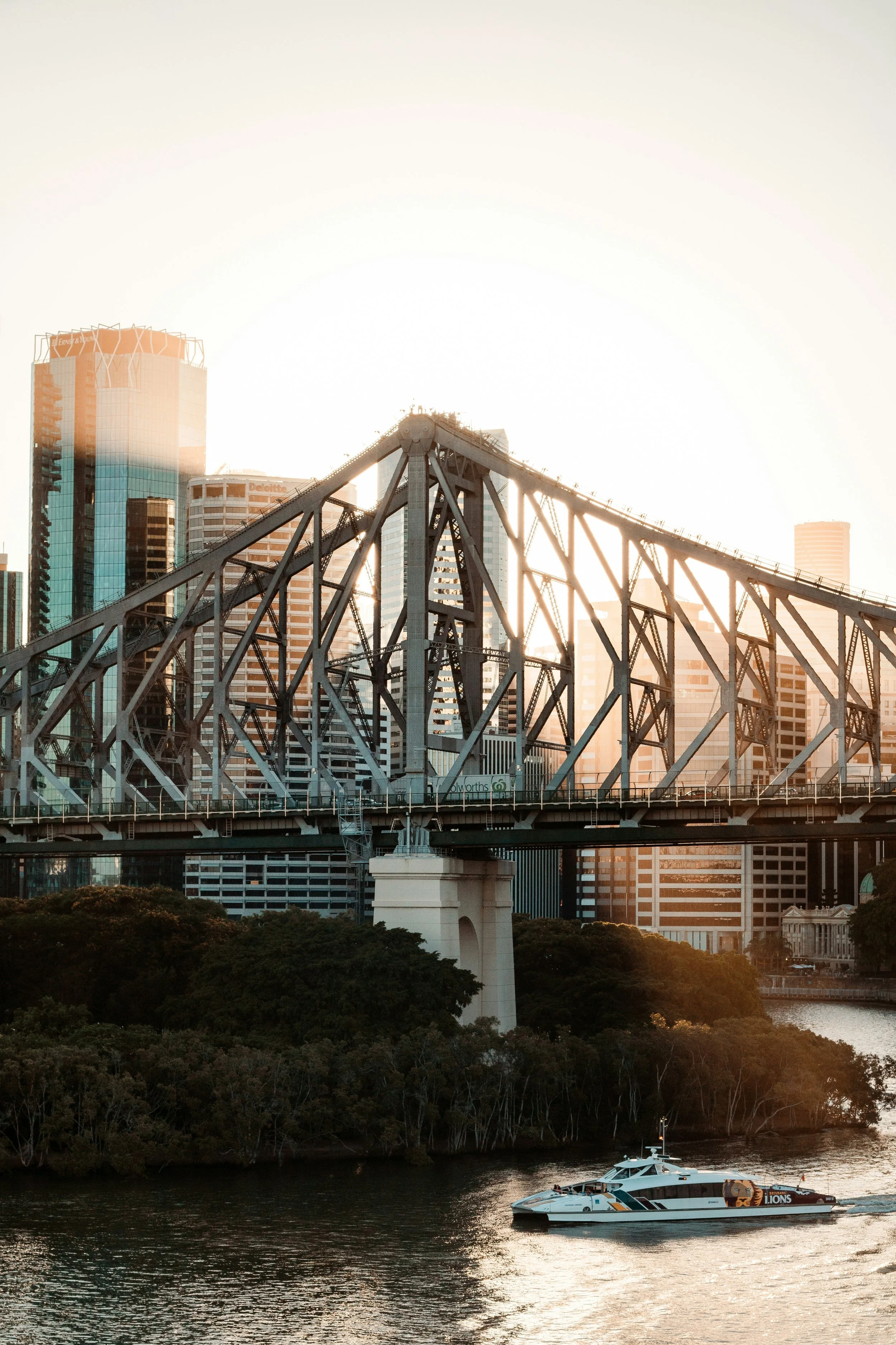 A cityscape with a steel bridge over water, tall modern buildings in the background, and a white boat on the water during sunset.