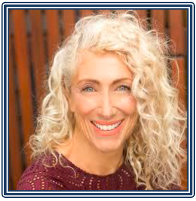 A woman with long, curly blonde hair smiling outdoors with a wooden fence in the background.