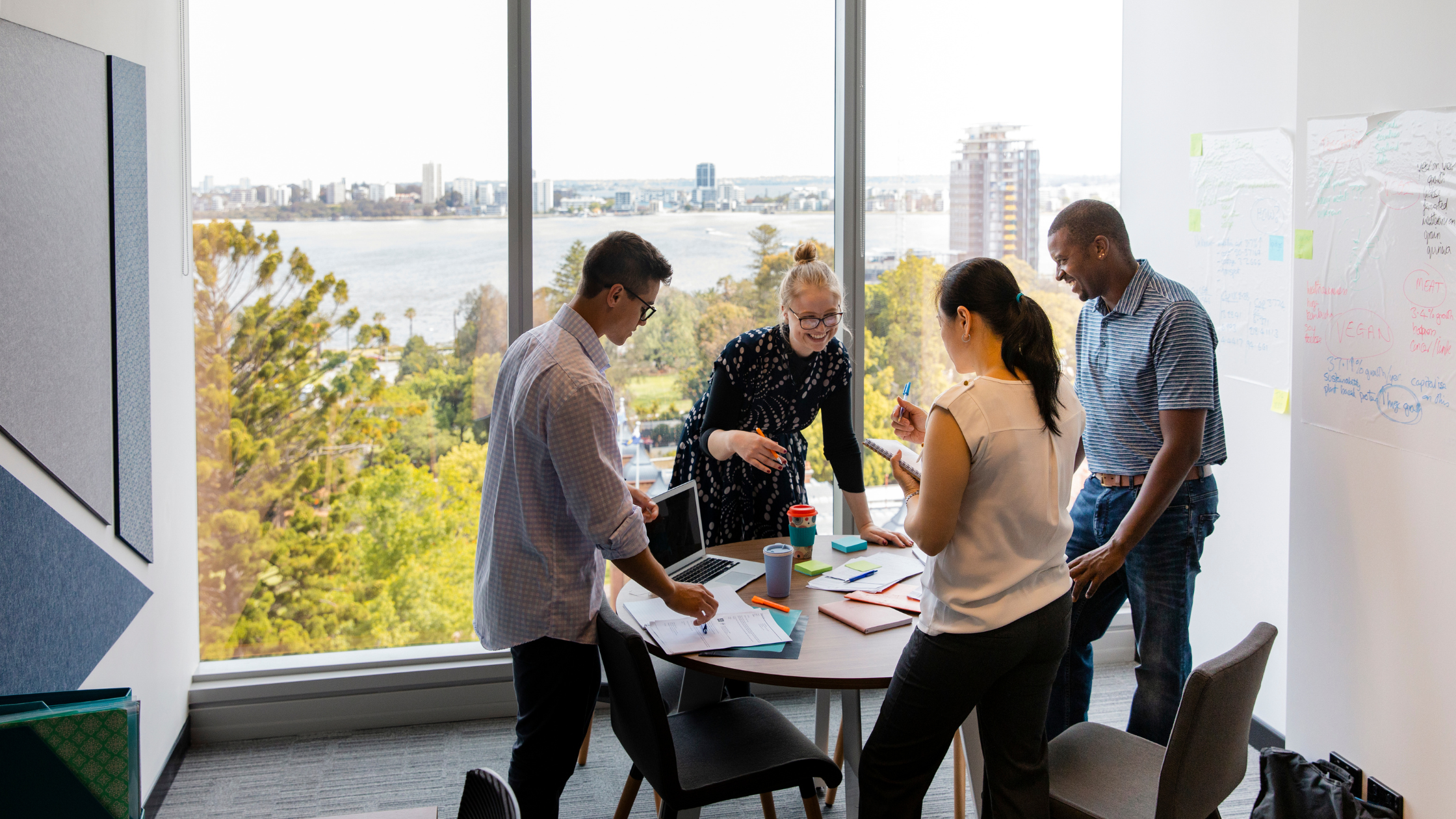 Five people in a bright office with large window discussing around a table with laptops, notebooks, and coffee cups.