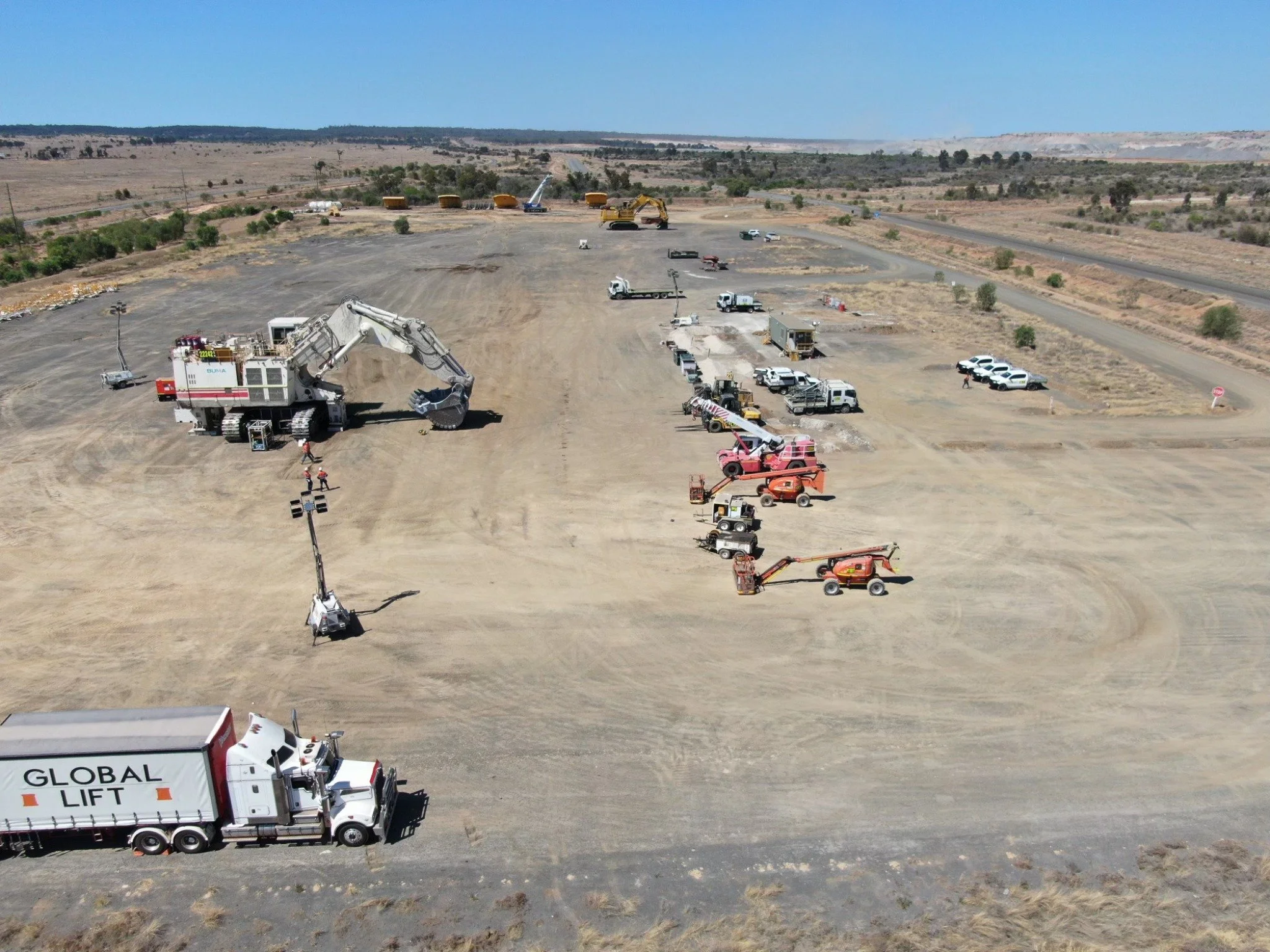 Construction site with heavy machinery, trucks, and vehicles on a large, flat dirt lot, with a clear blue sky and a rural landscape in the background.