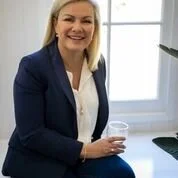 A woman with blonde hair smiling at a desk in an office with a large window in the background.