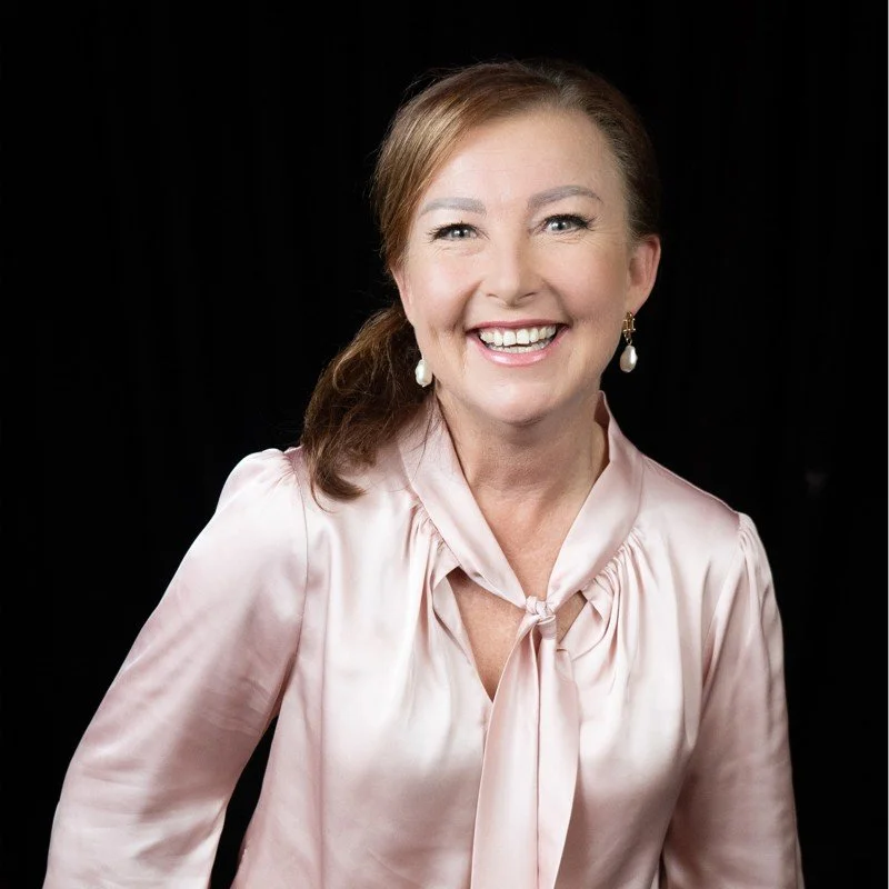 A woman with light brown hair tied back, smiling, wearing a pale pink satin blouse and pearl earrings, against a black background.