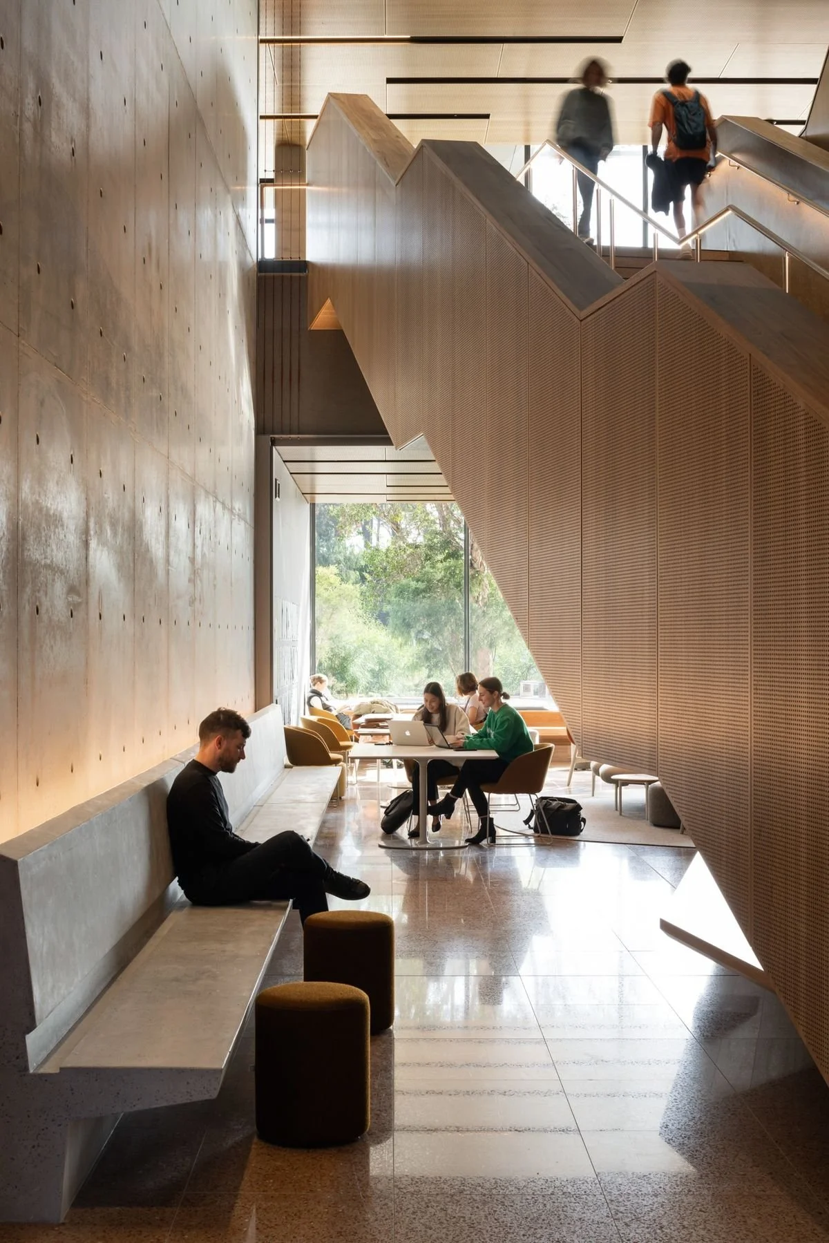 People sitting and working at a table in a modern, well-lit lobby or lounge area with a large window and architectural wooden staircase.