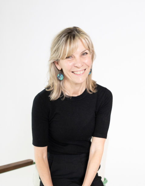 Portrait of a woman with wavy blonde hair, wearing a white top and turquoise earrings, smiling against a plain light background.