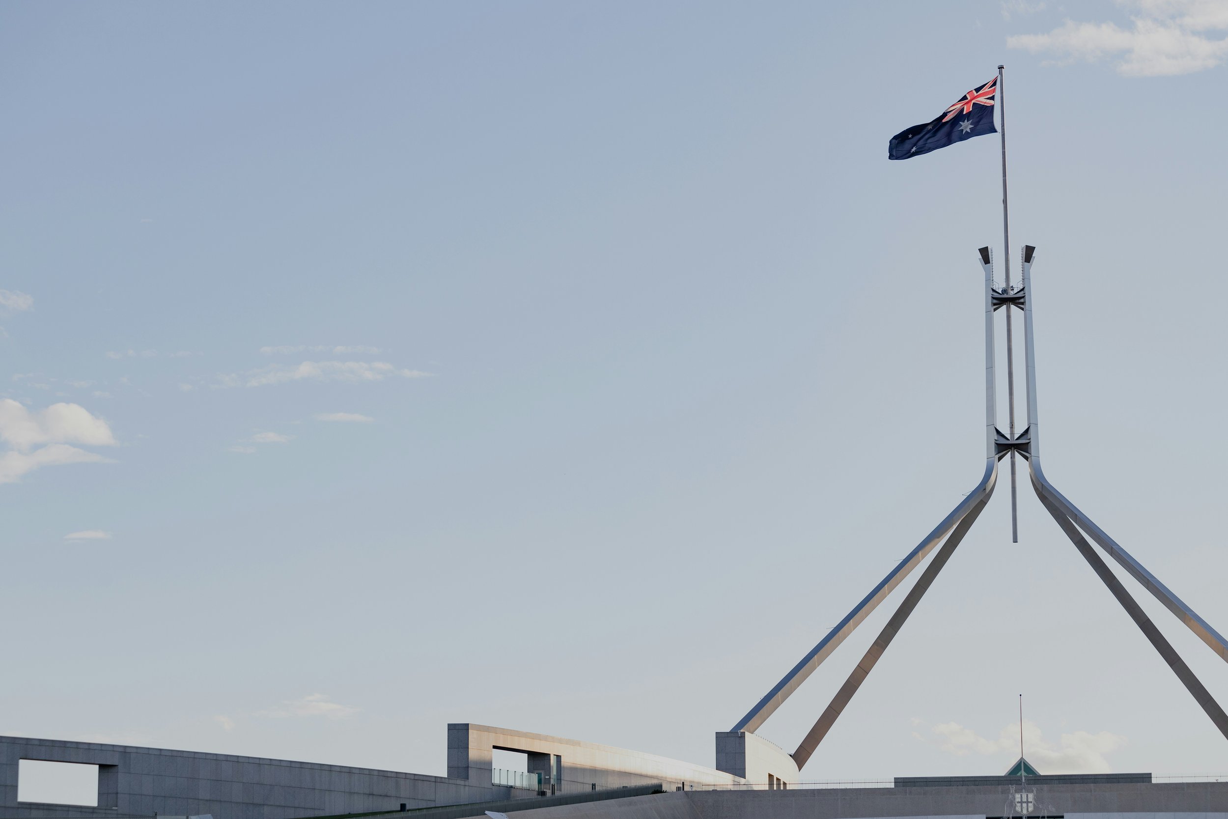 Australian flag flying from flagpole on a government building with a modern architectural design against a blue sky with wispy clouds.