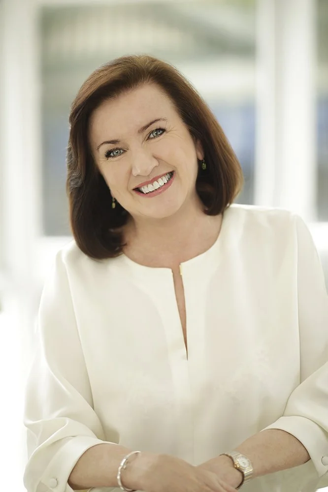 Portrait of a smiling middle-aged woman with brown hair, wearing a white blouse, standing indoors with blurred windows in the background.