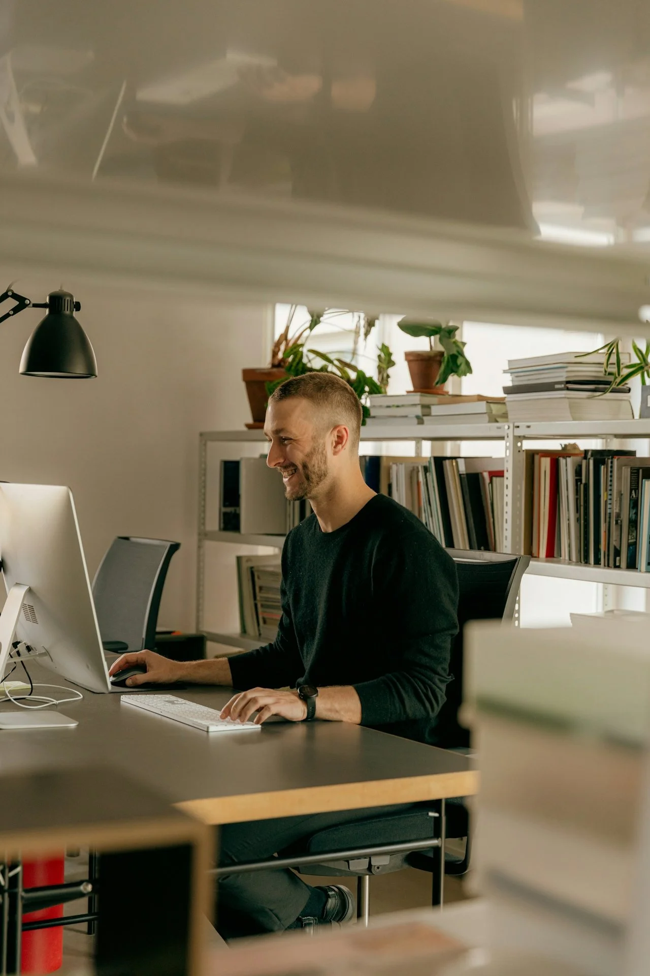 A man smiling while working at a computer in an office filled with books and potted plants.