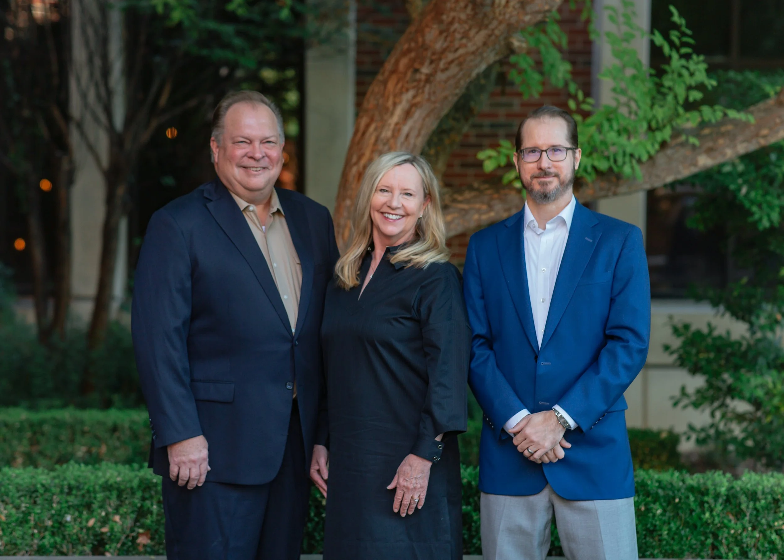 Three professionally dressed people standing outdoors in front of a tree with green foliage, smiling at the camera.
