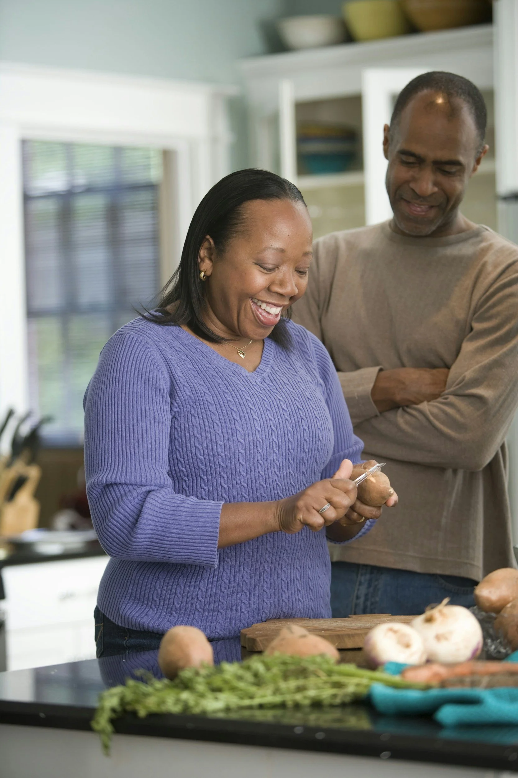 A woman in a purple sweater peeling potatoes while a man in a beige sweater stands nearby, smiling. They are in a kitchen with vegetables and green herbs on the counter.