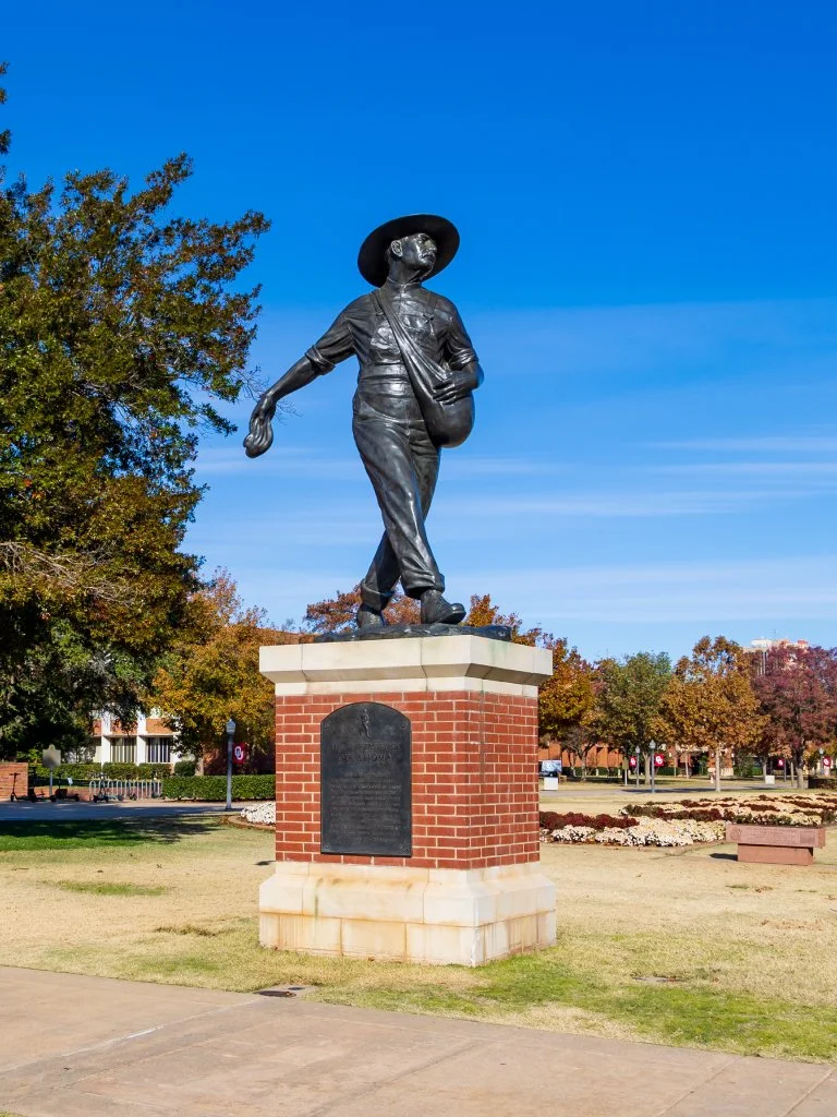 Bronze statue of the seed sower, wearing a wide-brimmed hat, walking with a bag slung over her shoulder, located on a brick pedestal in a park. University estate planning.