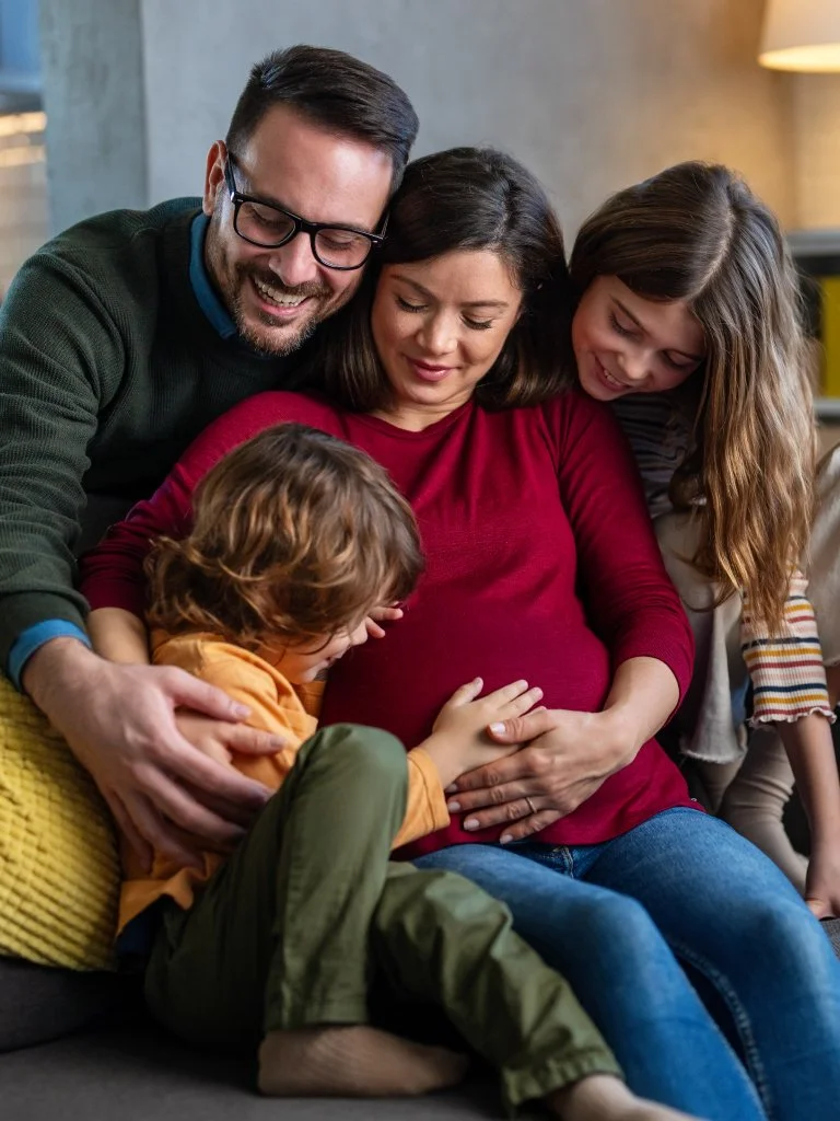A family of four, a pregnant woman, a man, a teenage girl, and a young boy, sitting closely together on a couch, happily sharing a moment filled with love and joy. Estate planning for blended families.