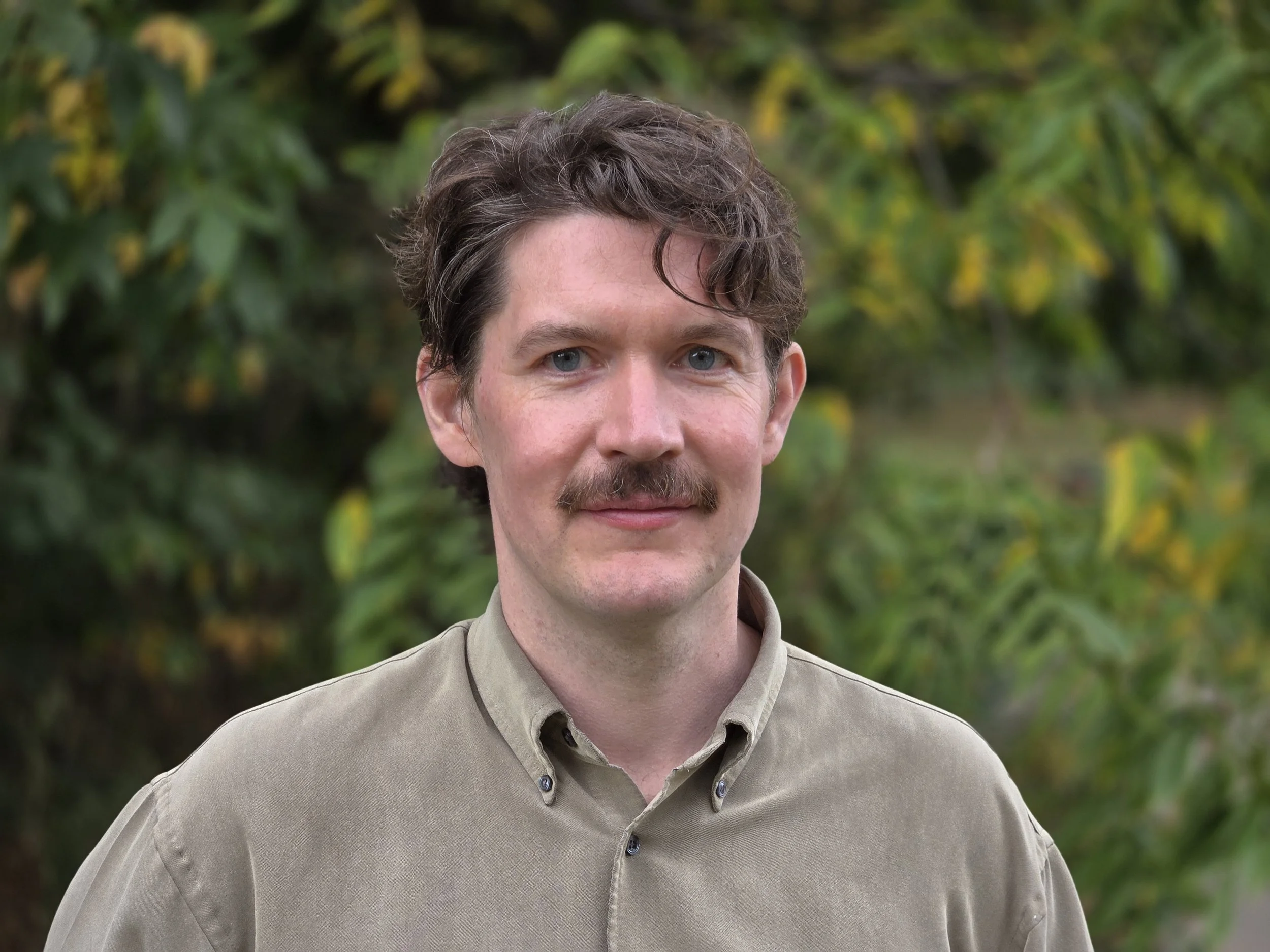 A man with light skin, blue eyes, and brown, slightly curly hair, wearing a beige button-up shirt, standing outdoors with a blurred background of green and yellow foliage.