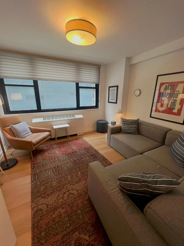 A living room with a large window, beige armchair, gray sofa with striped pillows, small side table, floor lamp, and framed art on the wall. There is a rug on light wood flooring, and a ceiling light fixture is on.