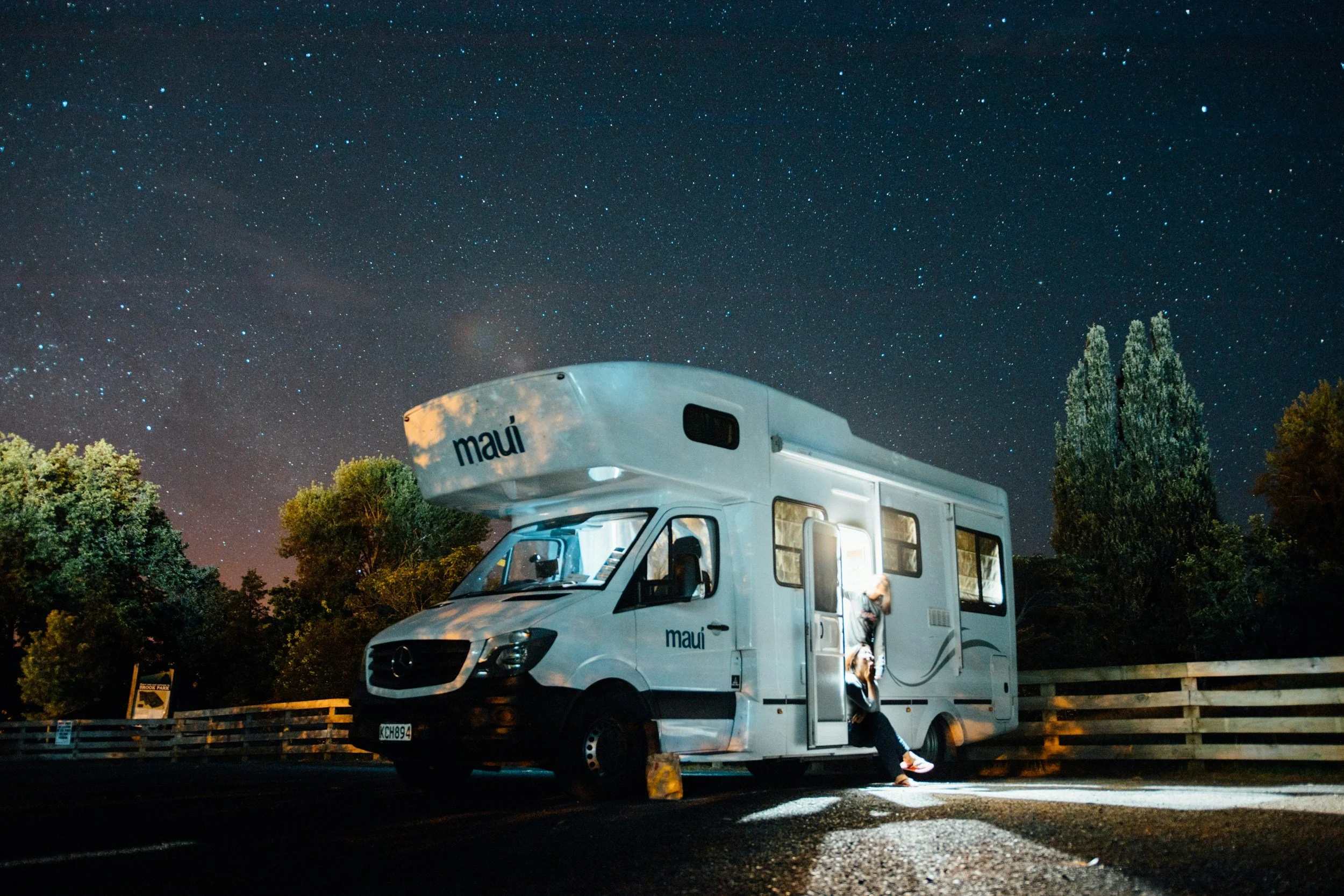 A person sitting outside a white camper van with the word 'maui' written on it, parked under a starry night sky with trees and a wooden fence in the background.