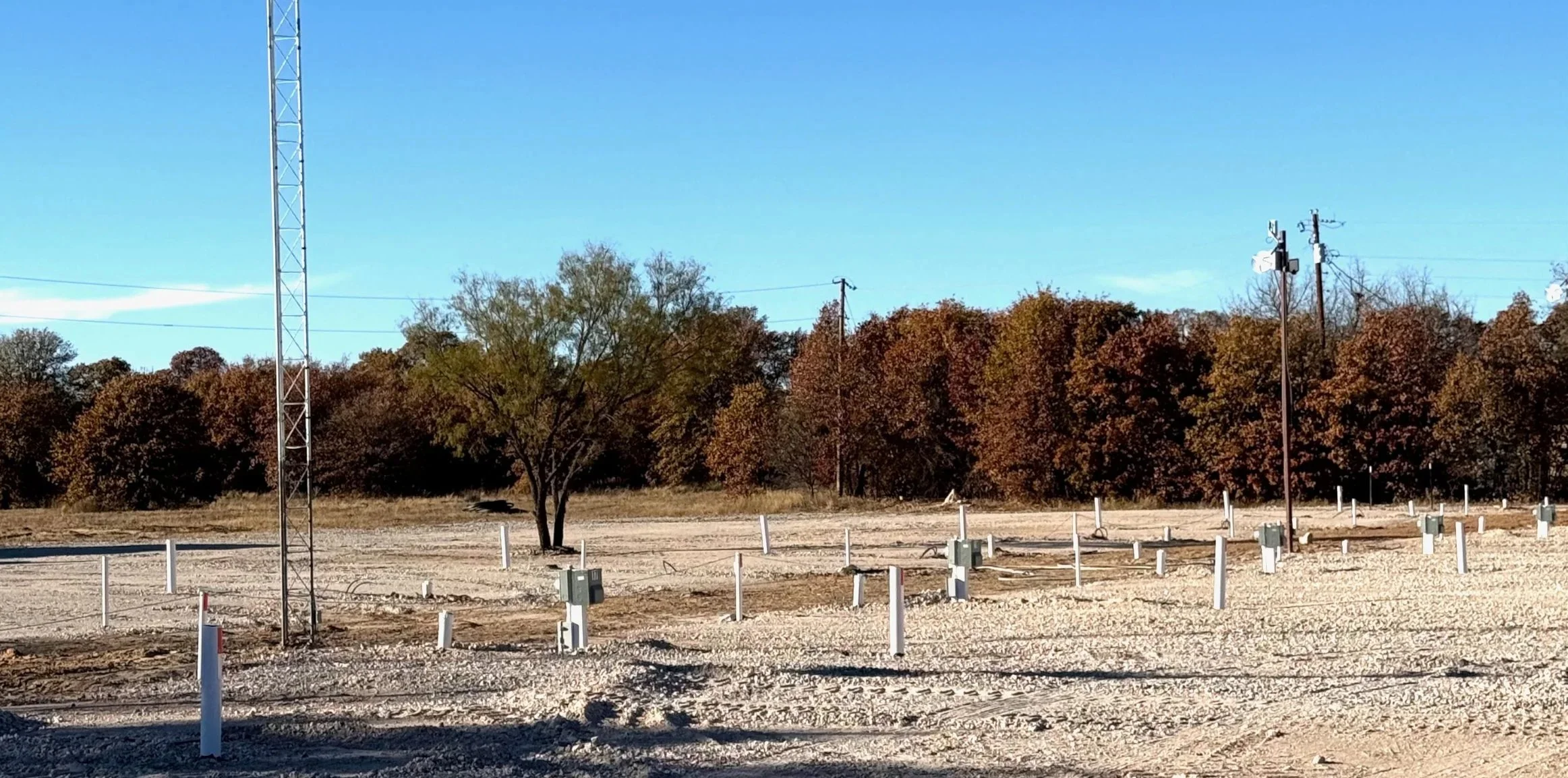 Open lot with multiple electrical boxes, poles, and utility wires, surrounded by trees with fall foliage under a clear blue sky.