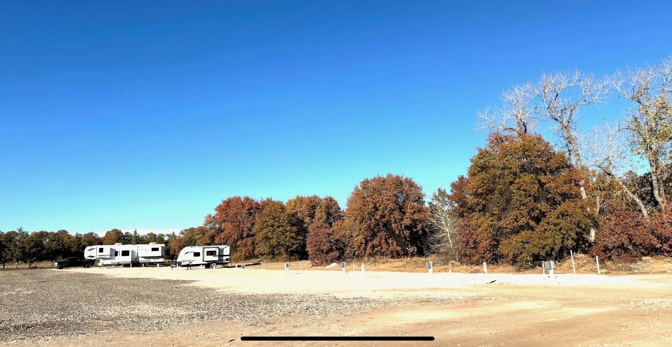 Camping site with three RV trailers parked in front of trees with colorful autumn leaves, under a clear blue sky.