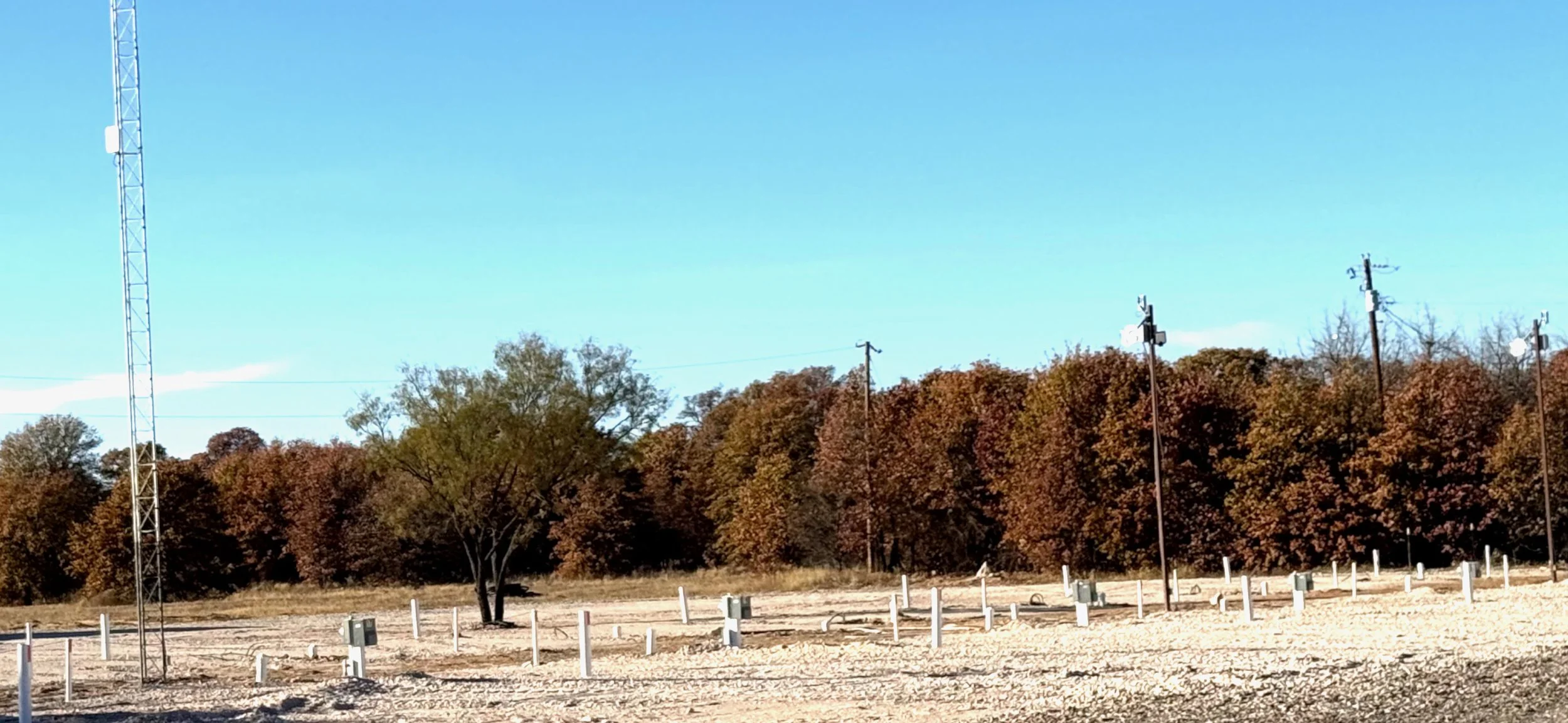 An outdoor landscape with a clear blue sky, trees with autumn foliage, and a gravel-covered area with utility poles and electrical boxes.
