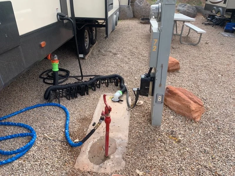 Camping area with in-ground water hook-up, electrical hookup, and blue hose on gravel ground near an RV, with picnic tables and rocks in background.