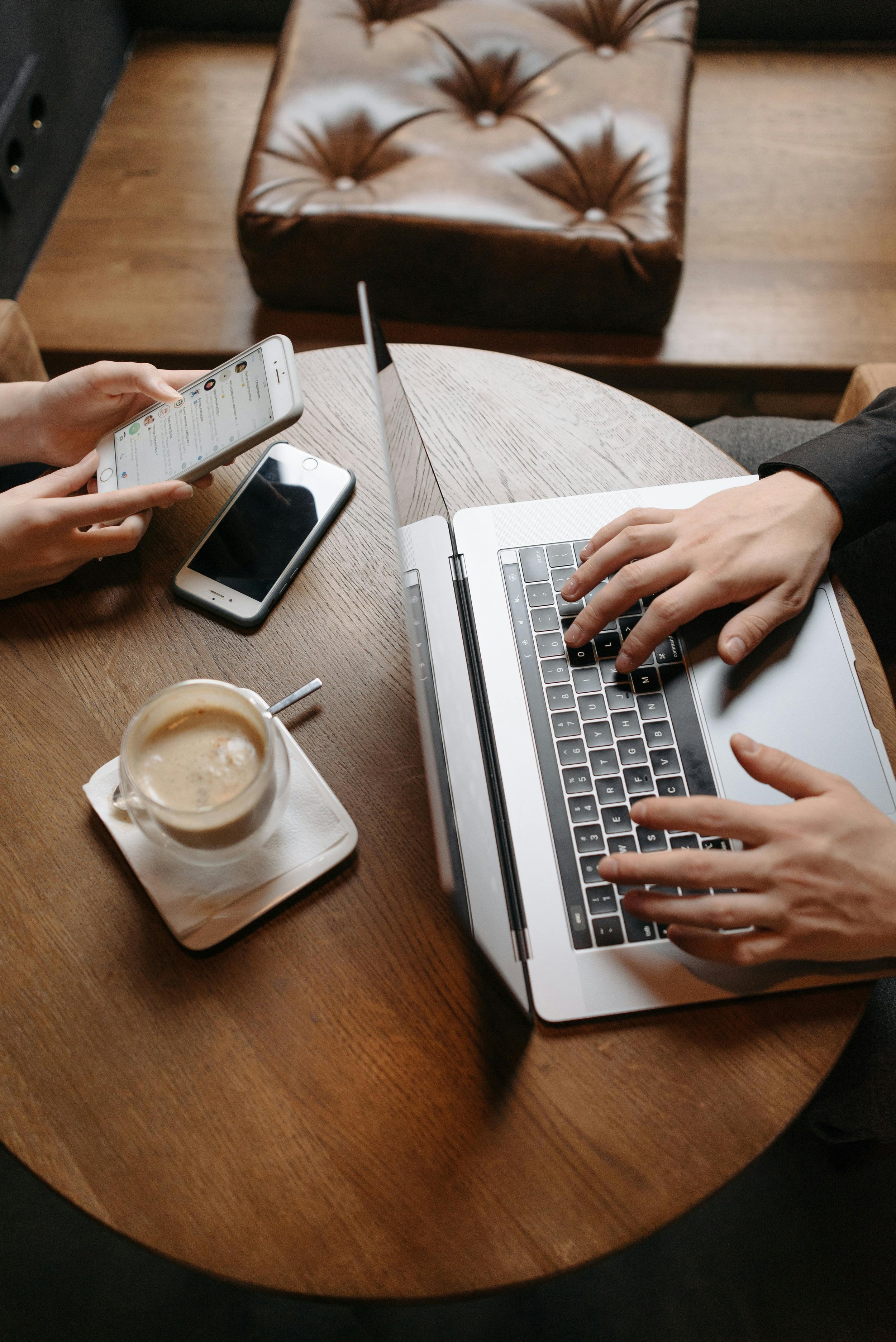 Two people working at a round wooden table with a laptop, two smartphones, and a coffee cup.