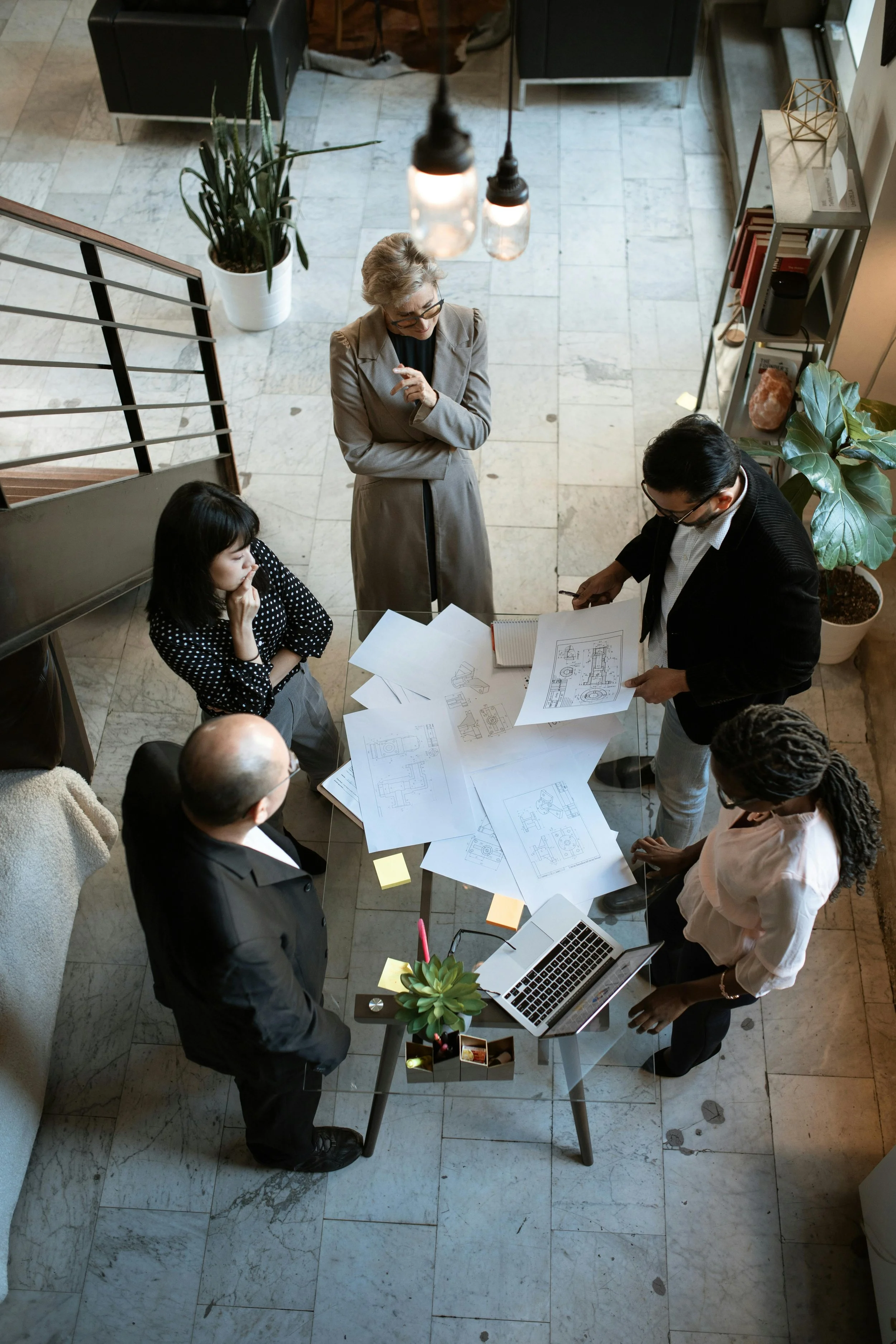 A group of five professionals in a meeting around a table with blueprints, documents, and a laptop in a well-lit office space.