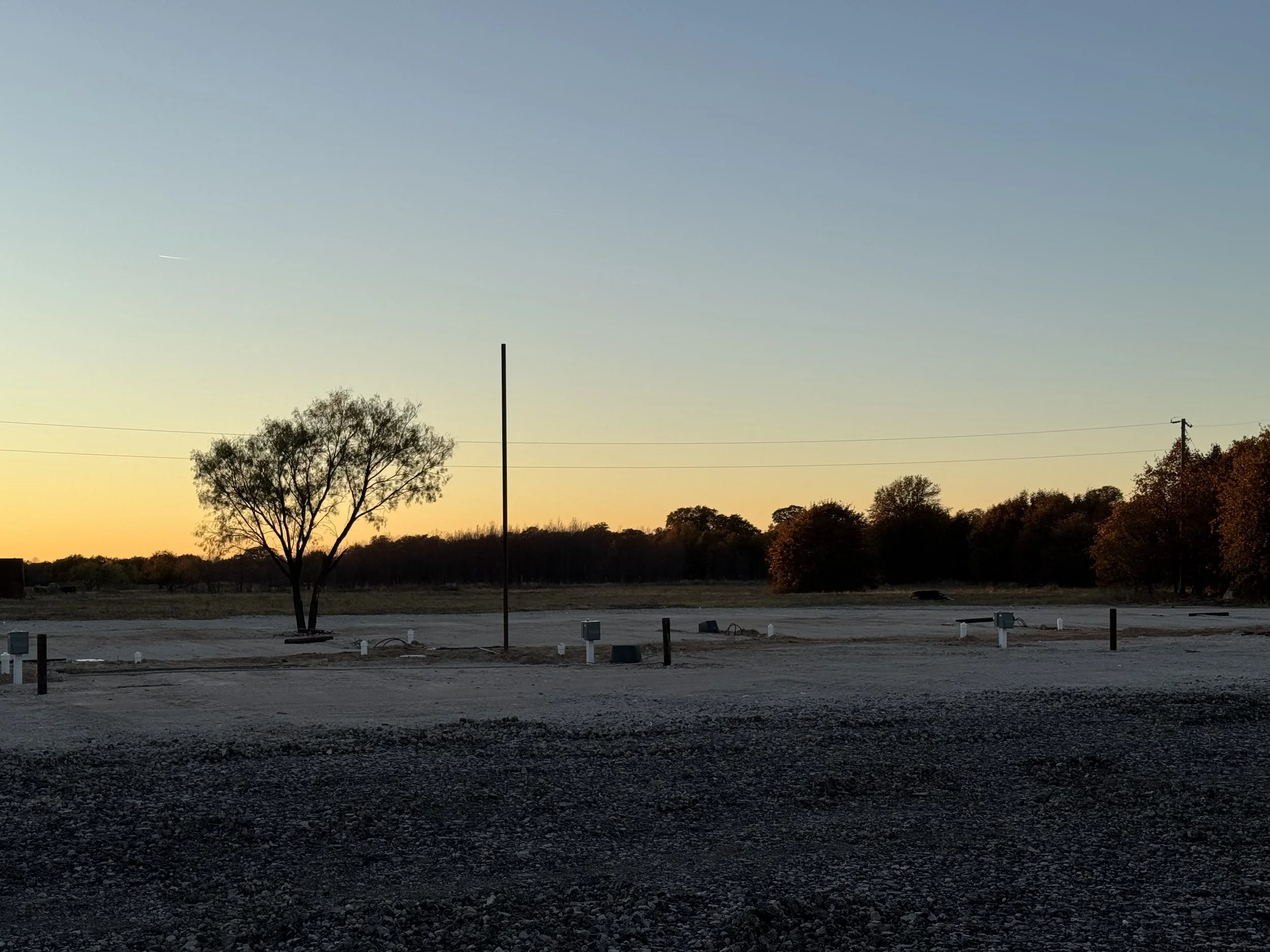 A rural landscape at sunset with a few trees, utility poles, and empty gravel parking spaces.
